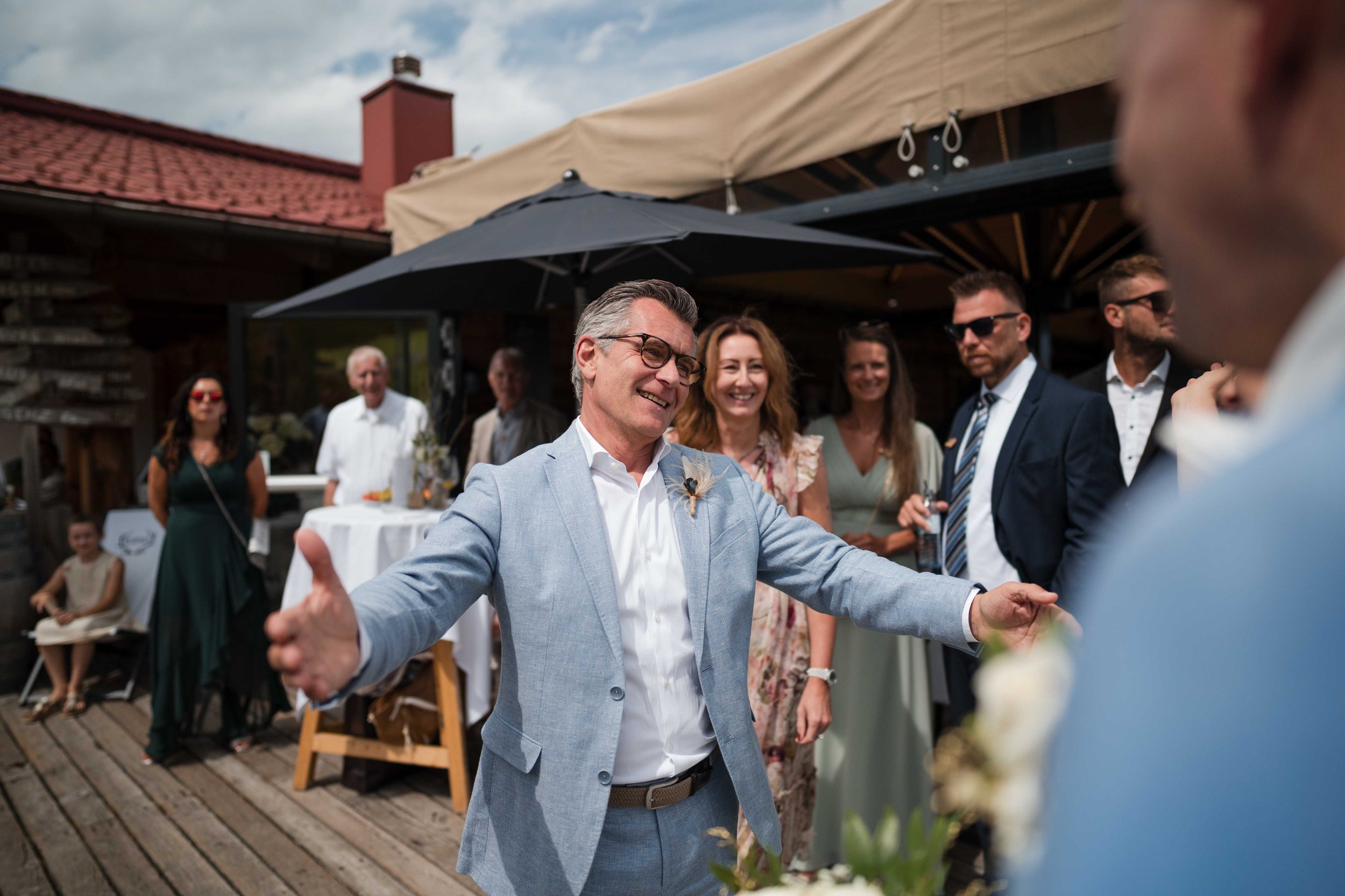 Hochzeitspaar bei einer Feier im Freien, umgeben von lachenden Gästen, mit einem Mann in einem hellblauen Anzug, der freundlich mit offenen Armen auf einen Bräutigam zugeht.