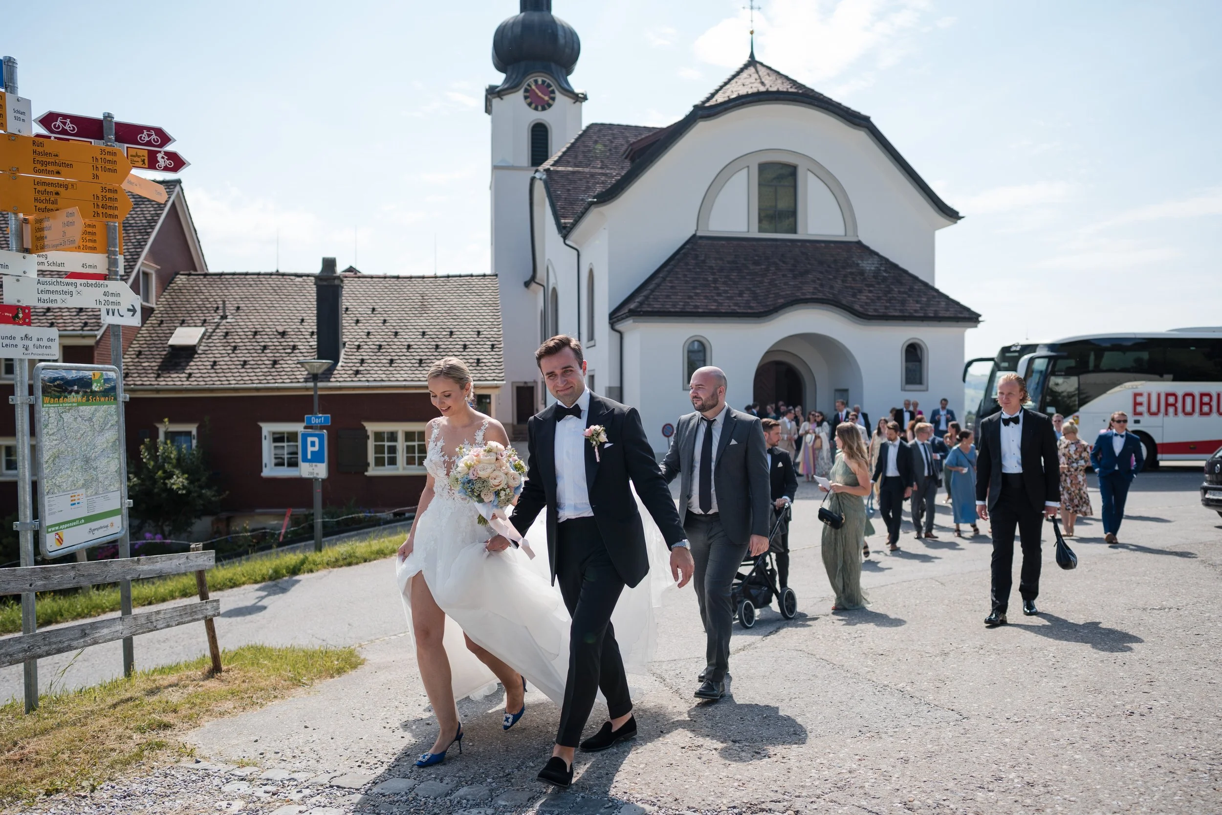 Ein Brautpaar in formeller Kleidung geht am Tag einer Hochzeit vor einer Kirche vorbei, umgeben von Gästen, auf einer öffentlichen Straße in einem kleinen Ort.