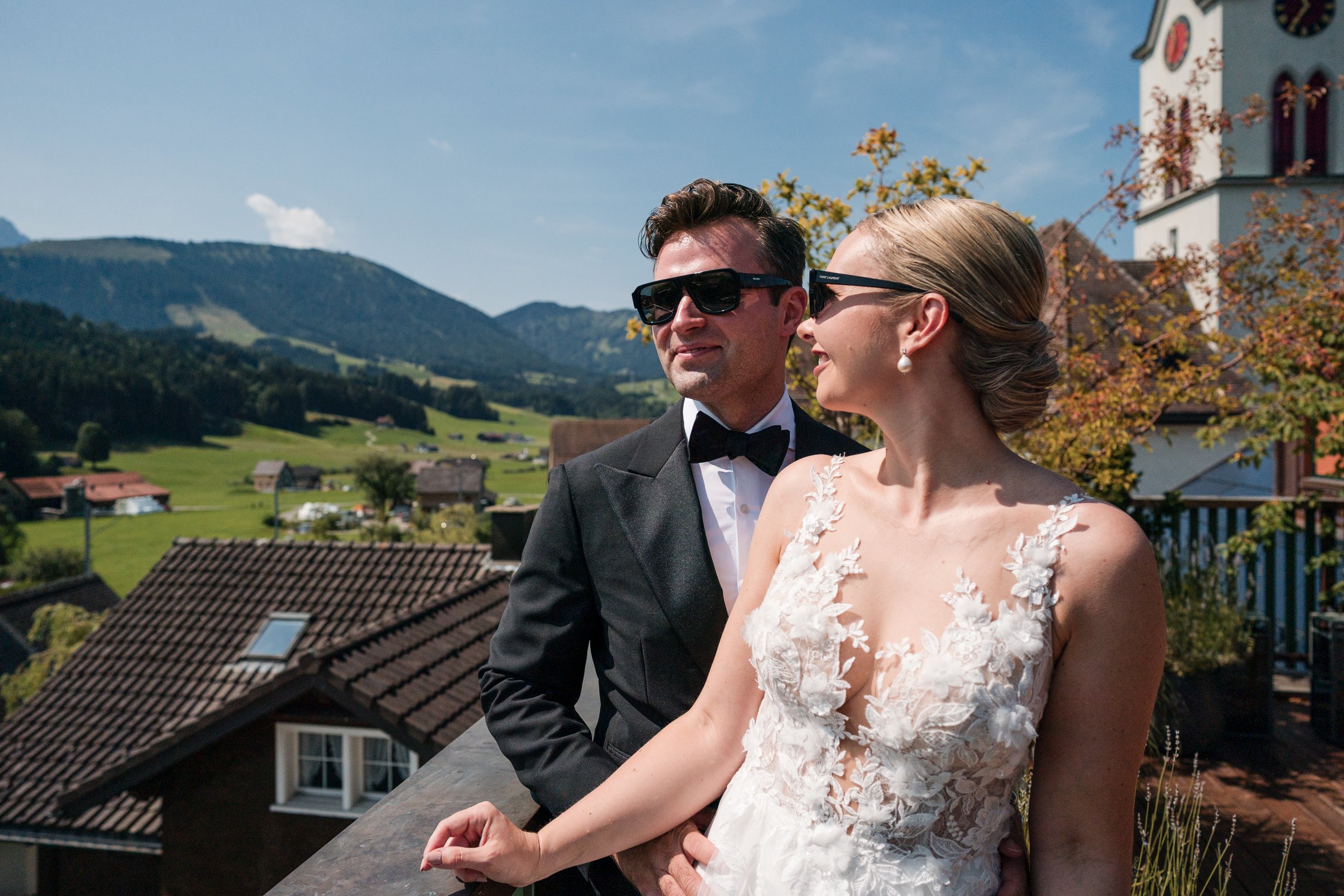Ein Brautpaar in formeller Kleidung, der Mann trägt einen schwarzen Anzug mit Fliege, die Frau ein weißes Hochzeitskleid mit floraler Spitze, beide tragen Sonnenbrillen, im Hintergrund eine ländliche Umgebung mit Hügeln und traditionellen Häusern, an