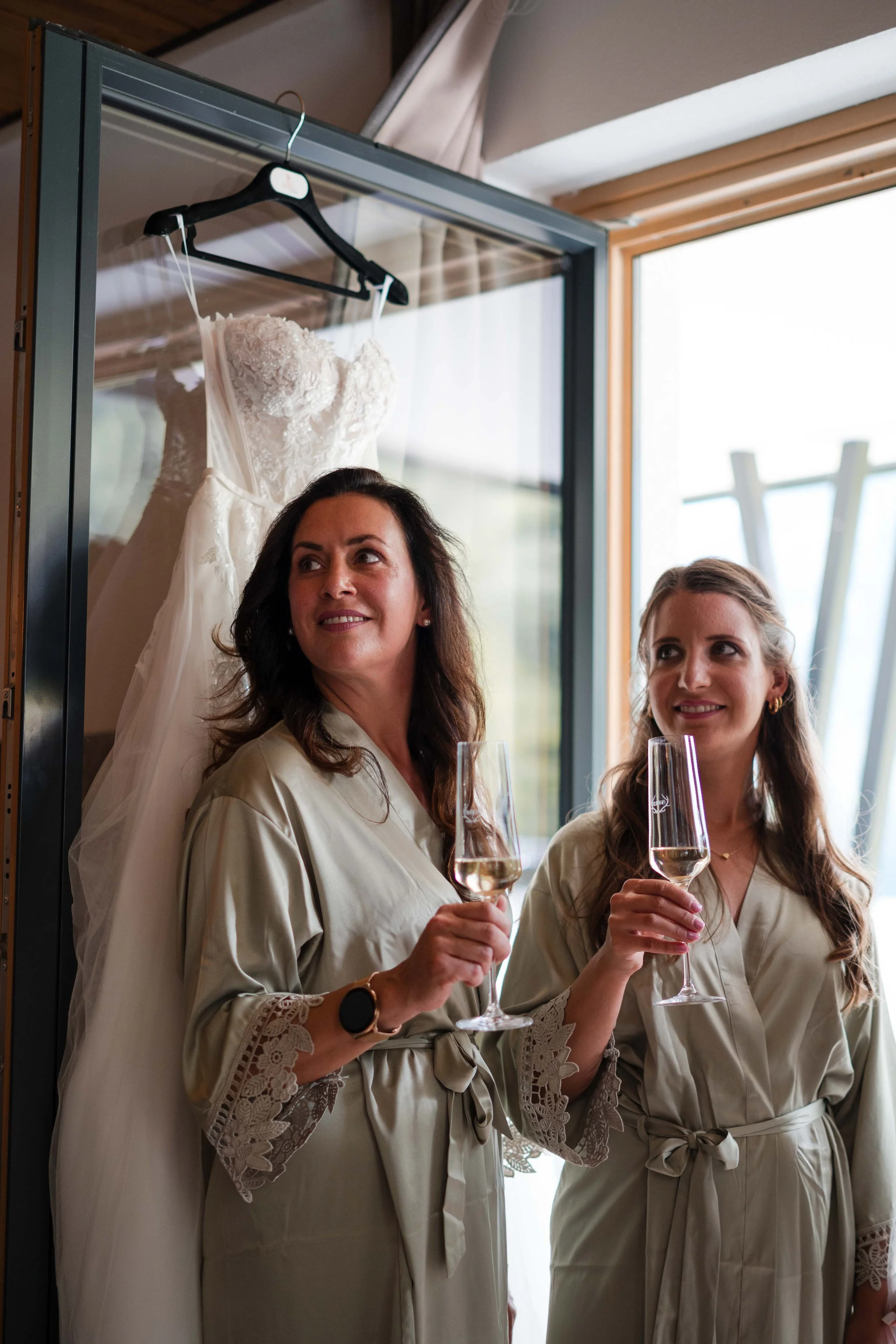 Zwei Frauen in Bademänteln mit Sektgläsern stehen im Zimmer, eine hat einen Hochzeitskleid auf einer Hängegarderobe im Hintergrund.