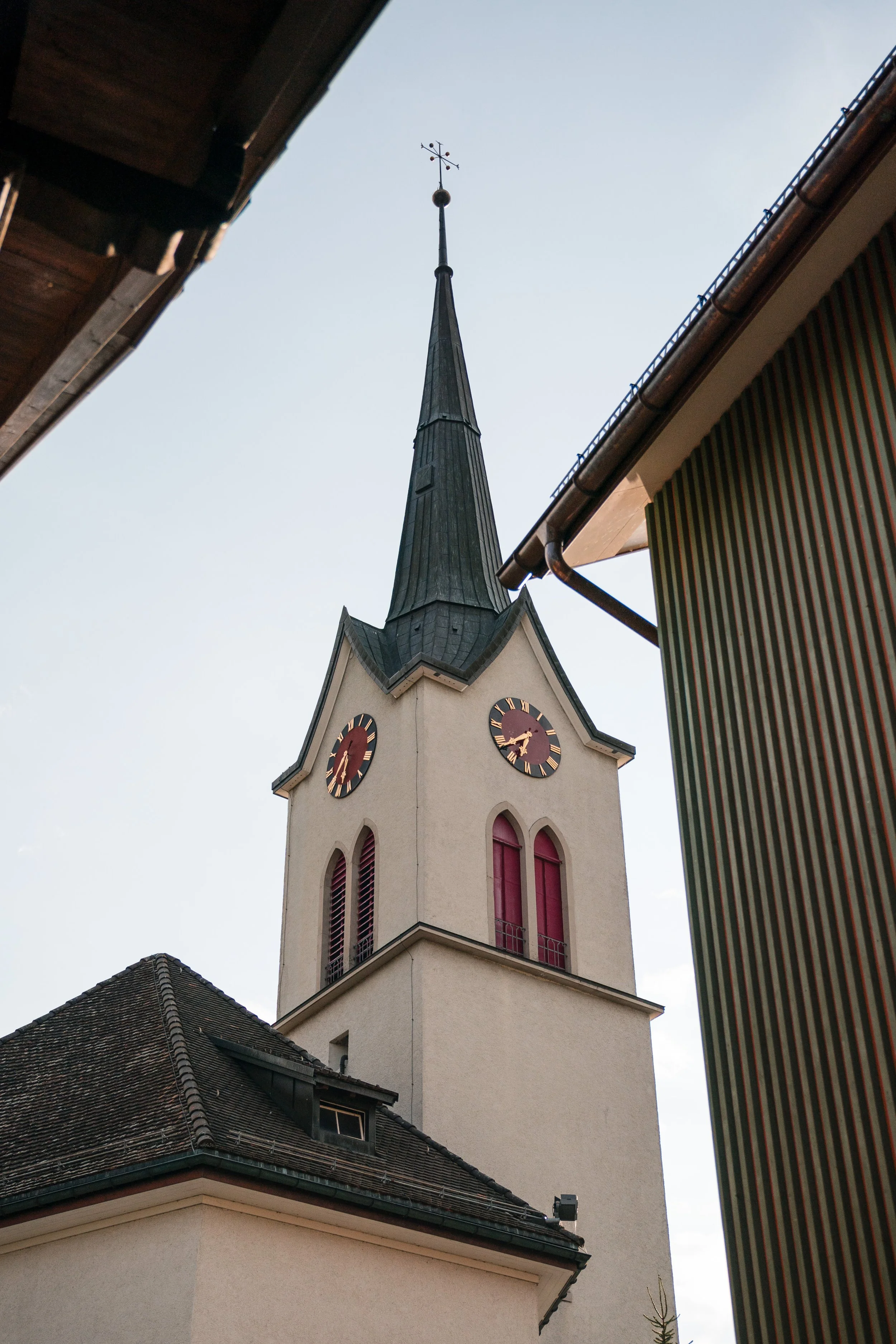 Blick auf den Kirchturm einer Kirche mit zwei Uhren, spitze Dach und Wetterhahn, umgeben von Gebäuden.
