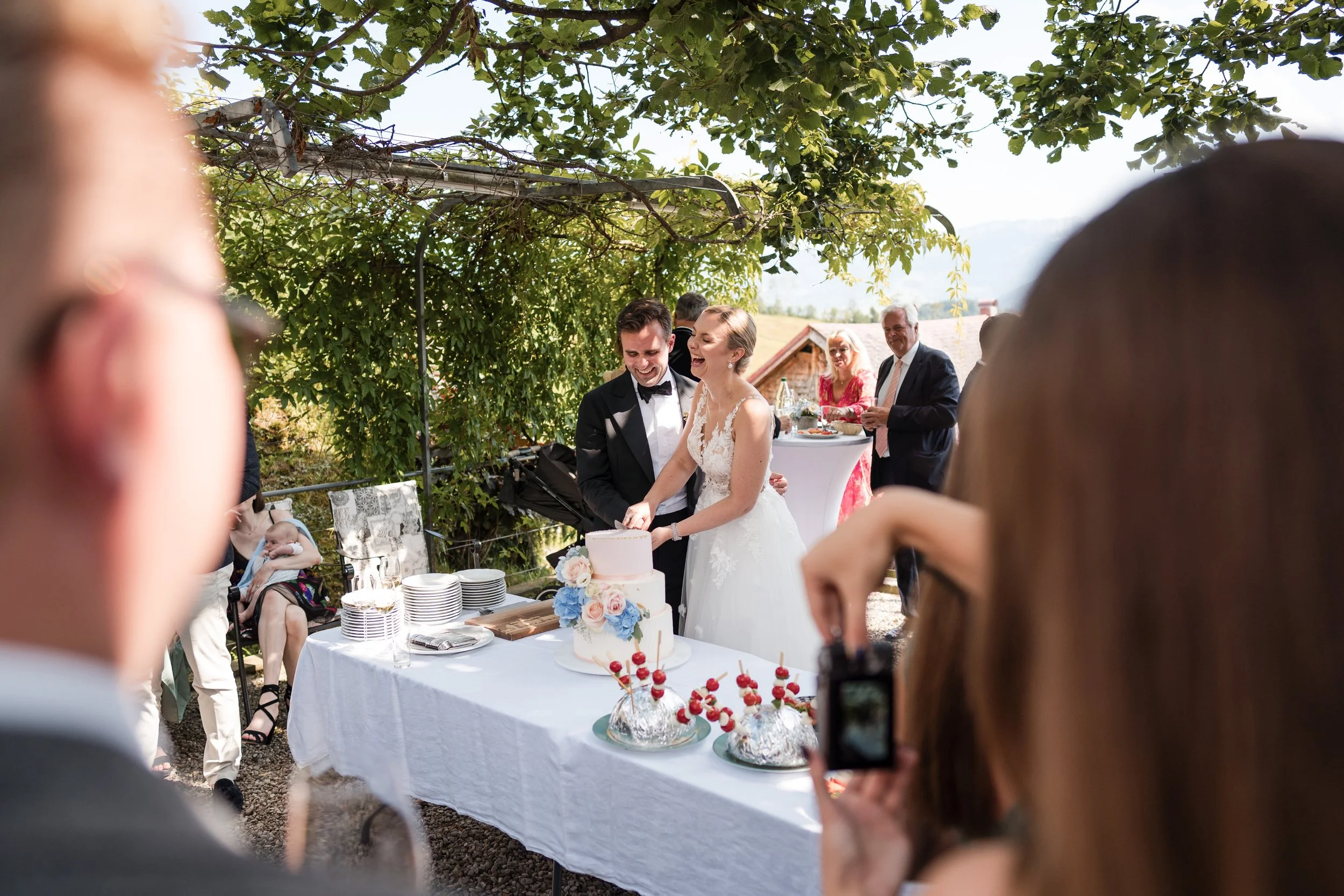 Ein Brautpaar schneidet eine Hochzeitstorte beim Empfang im Freien, umgeben von Gästen und einem grünen Baumdach.