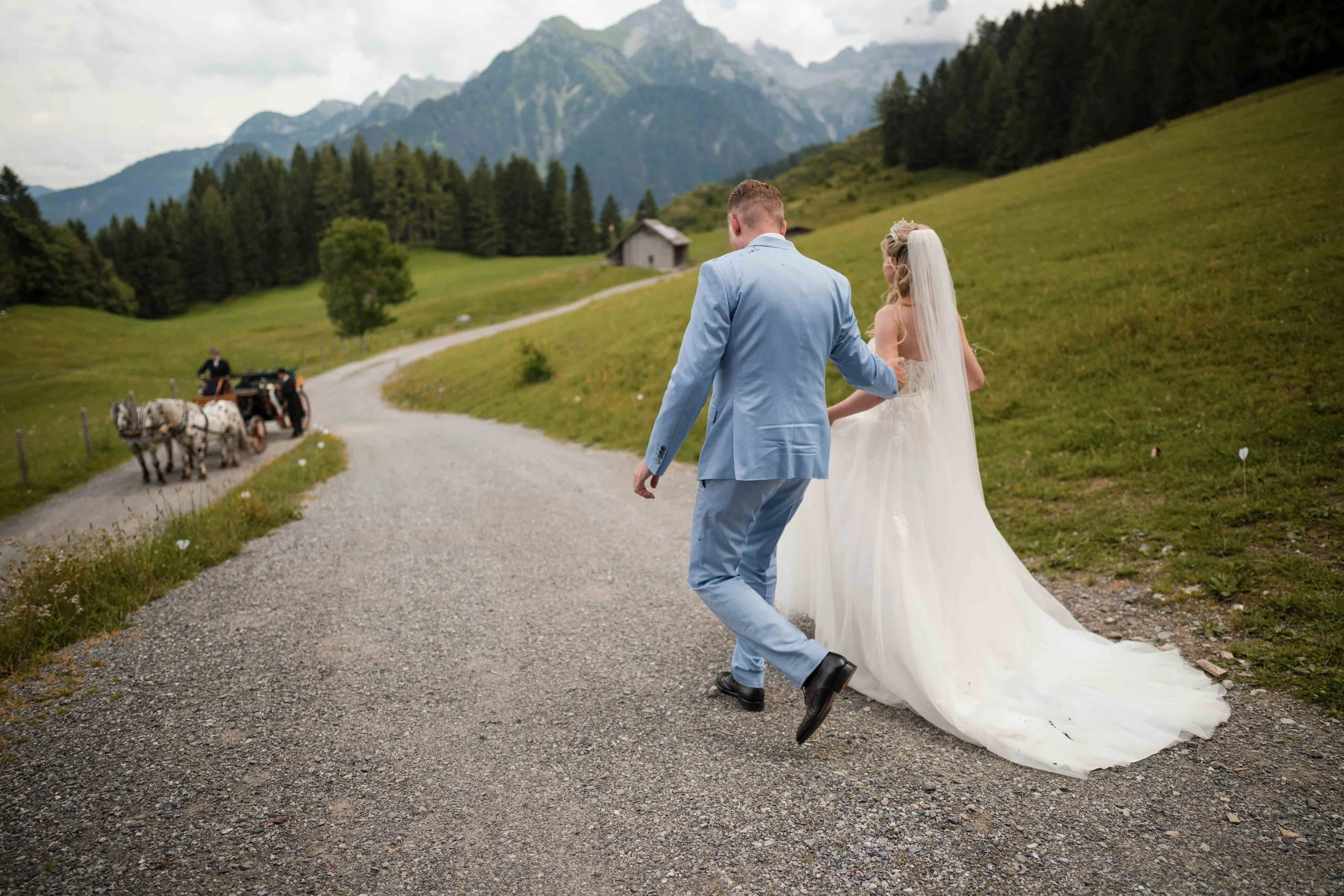 Ein Brautpaar läuft Hand in Hand auf schmalen Kiesweg in einer grünen, hügeligen Landschaft mit Bergen im Hintergrund. Der Bräutigam trägt einen hellblauen Anzug, die Braut ein weißes Hochzeitskleid mit Schleier. In der Ferne zieht eine Kutsche mit P