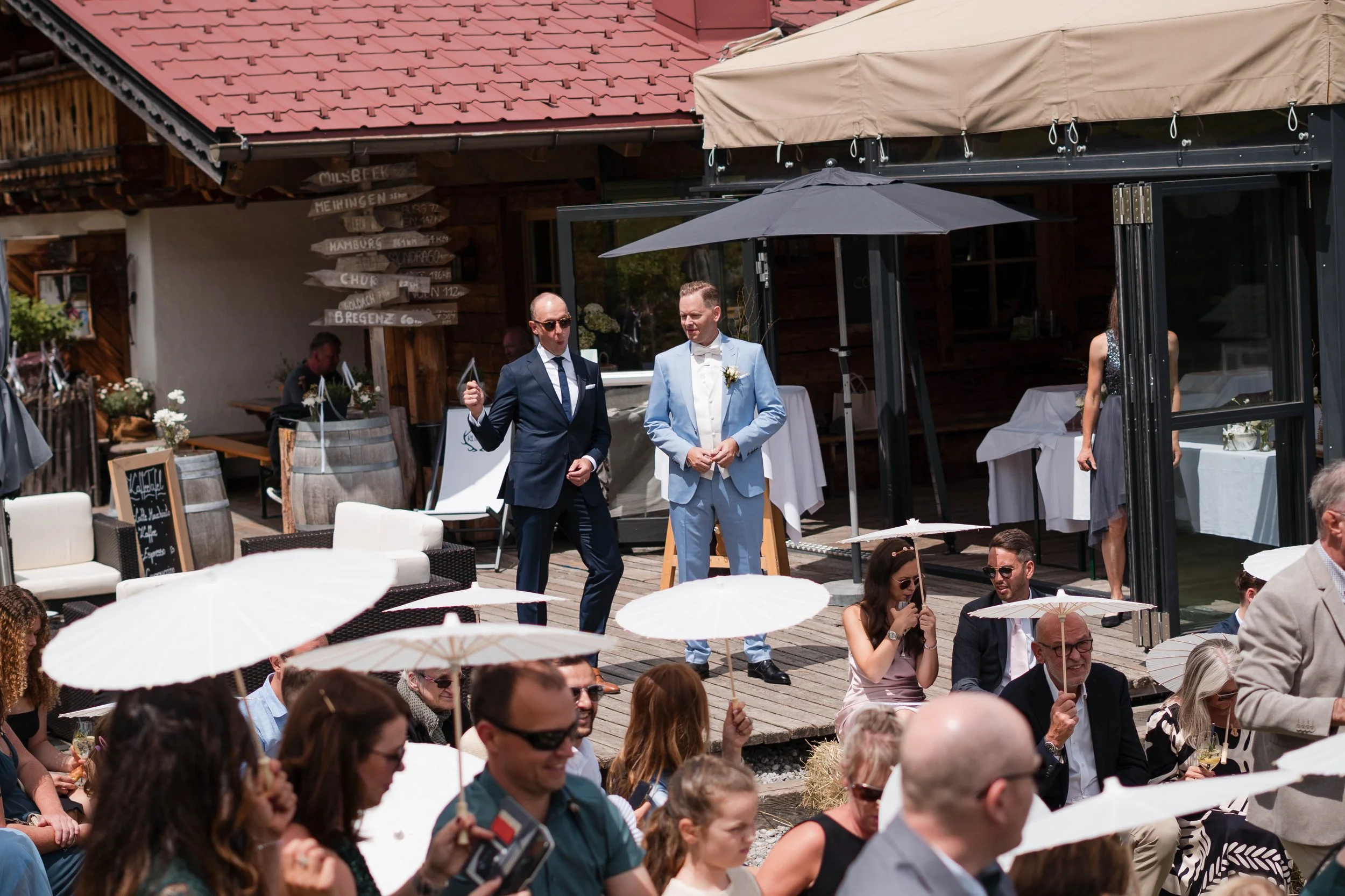 Hochzeit im Freien mit Gästen, die Unterhemden und Sonnenschirme tragen, ein Bräutigam in hellblauer Anzug, ein Mann in dunklem Anzug, und eine Frau in einem kleinen Kleid bei einer Zeremonie auf einem Holzboden. Im Hintergrund sind Bars und Dekorati