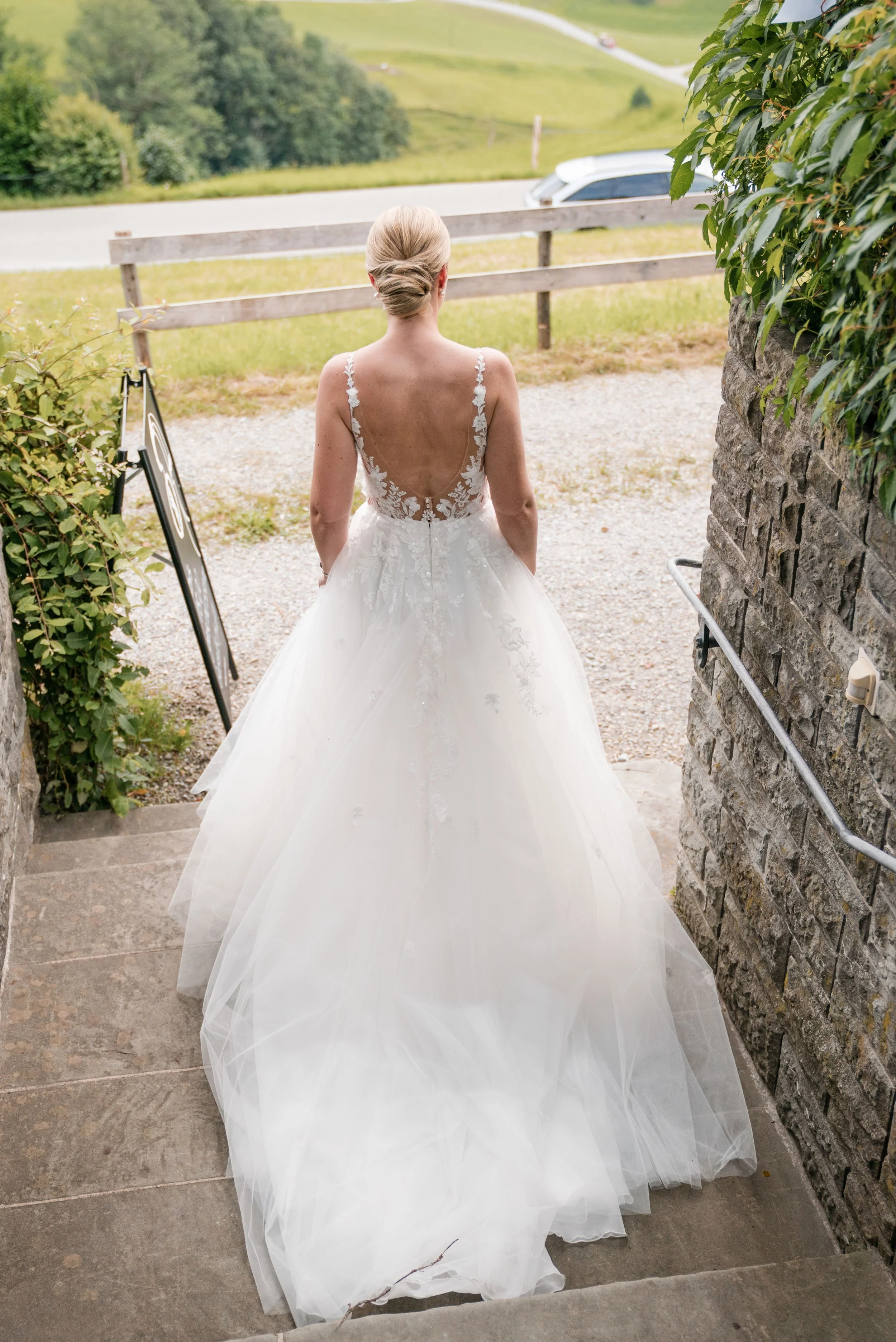 Braut in einem weißen Hochzeitskleid mit tiefem Rückenausschnitt steht auf Treppen vor einer ländlichen Szenerie mit grünen Hügeln, Bäumen und einem Weg.