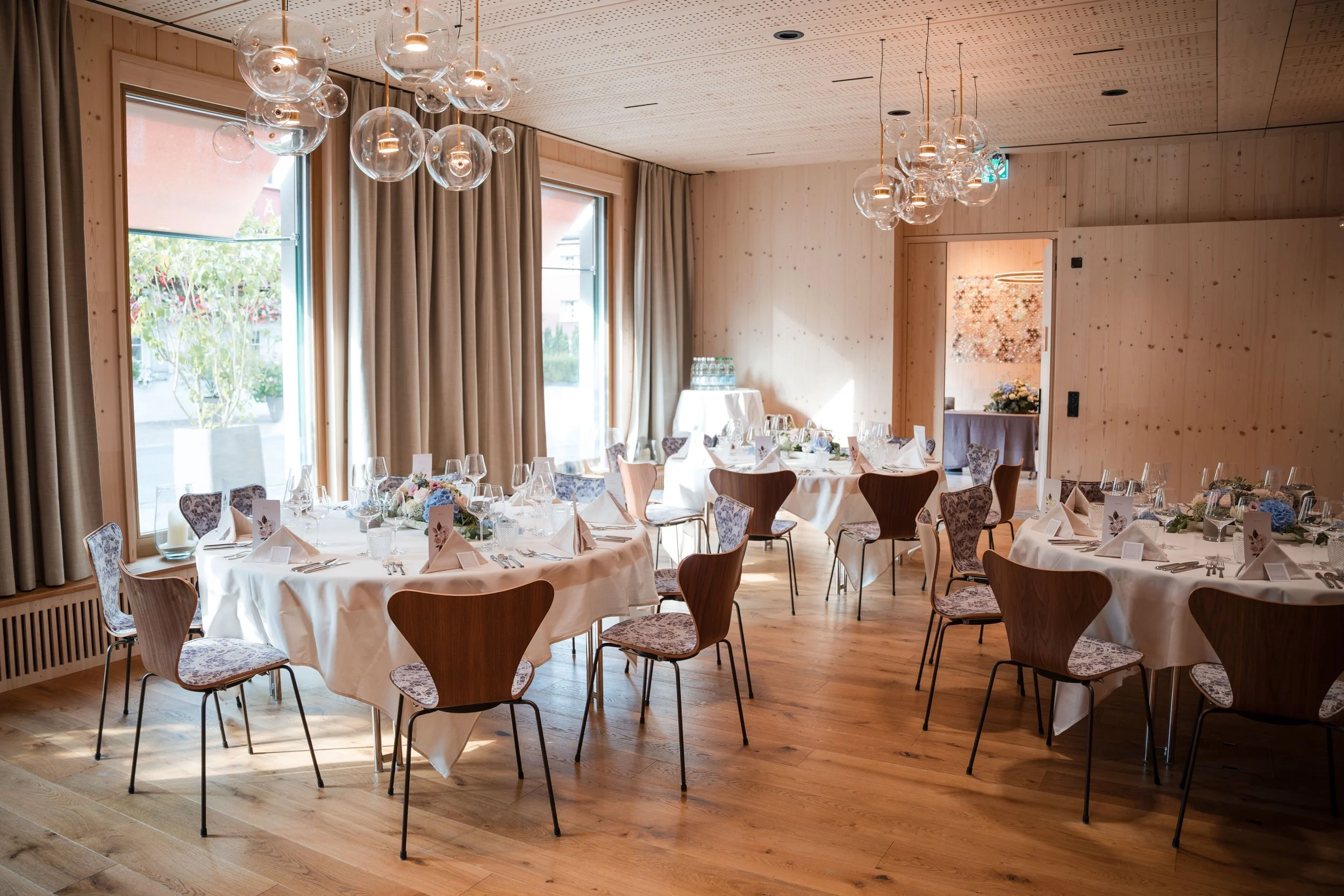 Ein schön gedeckter Veranstaltungsraum mit mehreren runden Tischen, weißen Tischtüchern, Blumenarrangements und Besteck, mit großen Fenstern und Holzinterieur.