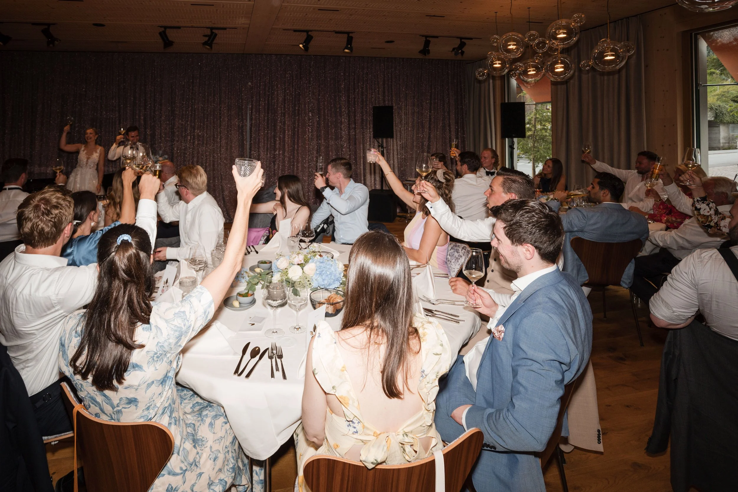 Menschen bei einer Feier sitzen an Tischen, trinken, lachen und heben ihre Gläser bei einem Fest im Innenraum.