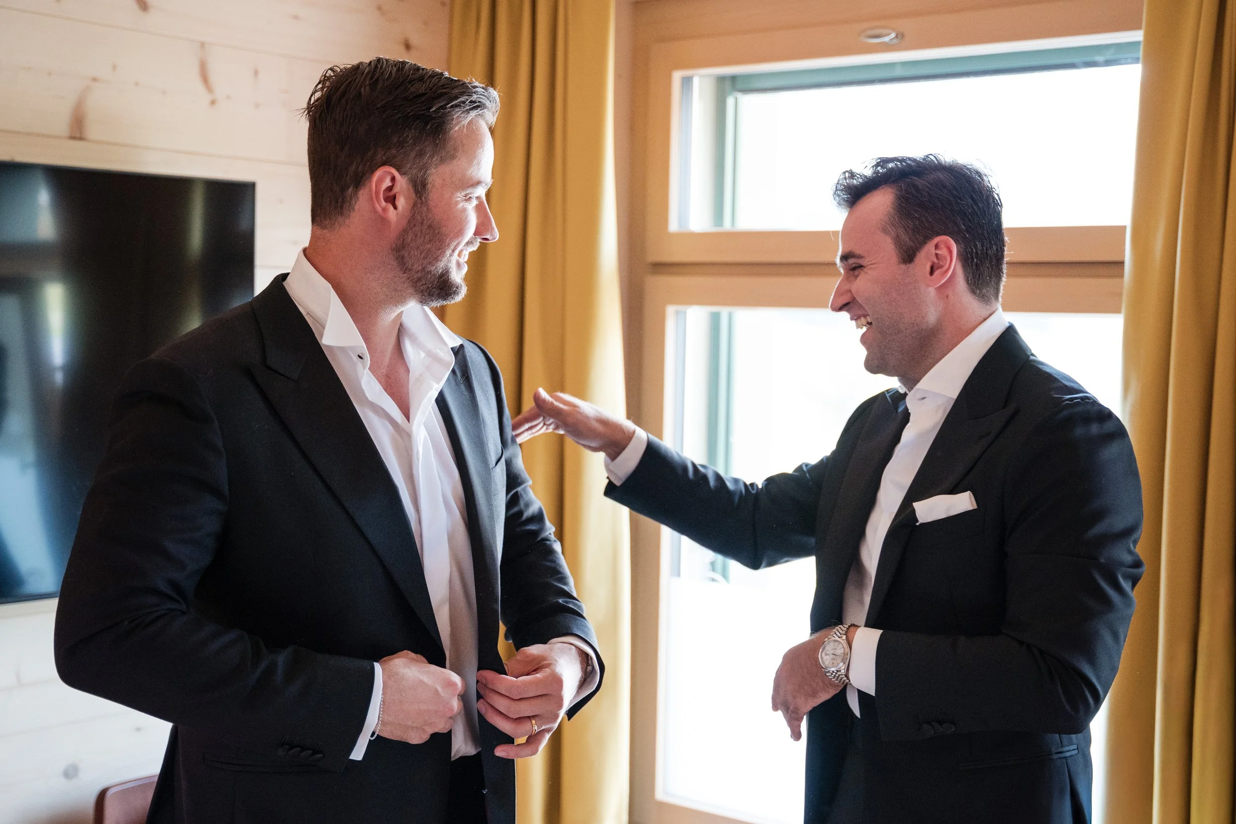 Zwei Männer in schwarzen Anzügen lachen und unterhalten sich in einem Raum mit großen Fenstern und gelben Vorhängen.