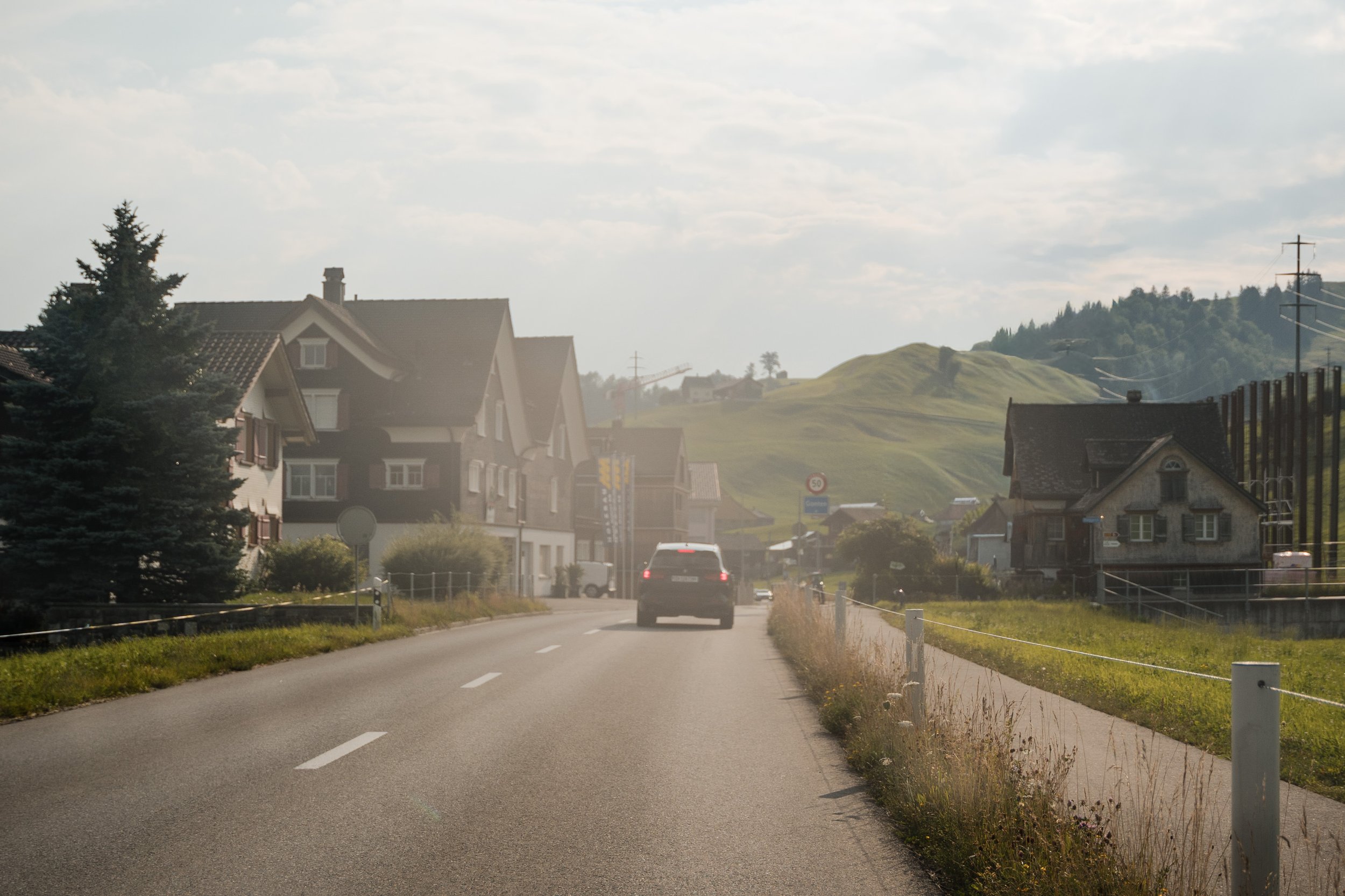 Eine Straße in einer ländlichen Gegend mit Häusern auf beiden Seiten, grüne Hügel im Hintergrund, ein Auto fährt in Richtung Berge, bewölkter Himmel.
