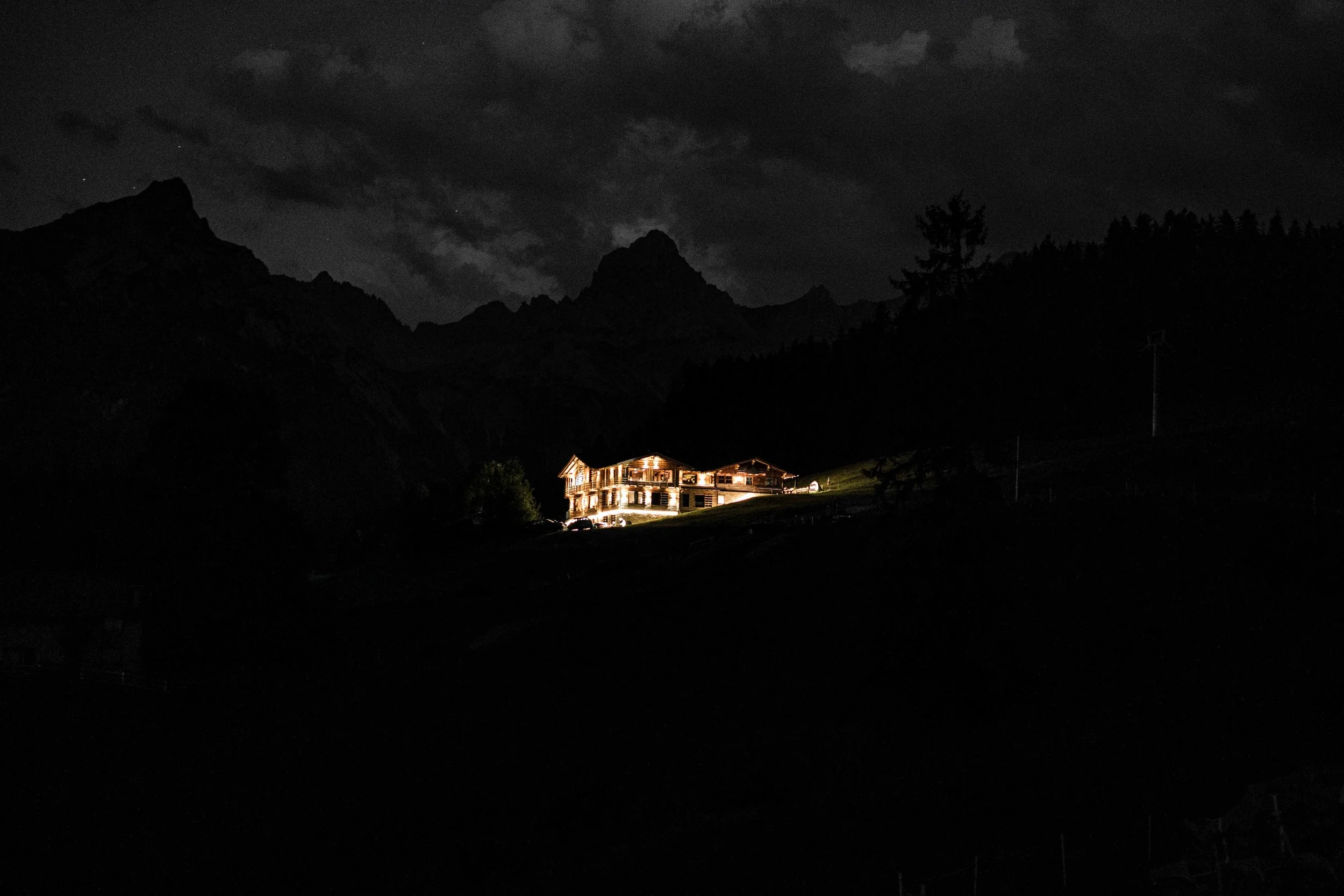 Ein beleuchtetes Haus auf einer Wiese in einer bergigen Landschaft bei Nacht, dunkle Wolken am Himmel, im Hintergrund Berge