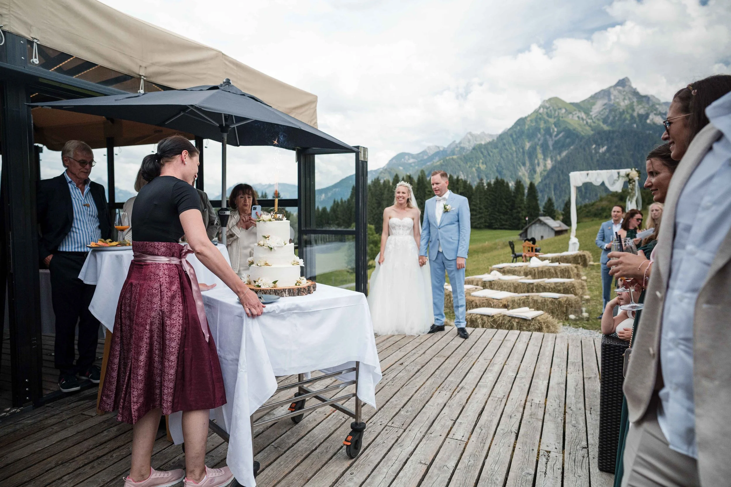 Ein Brautpaar bei ihrer Hochzeit im Freien, umgeben von Gästen, mit einer Hochzeitstorte auf einem Tisch. Die Szenerie ist von Bergen und einer grünen Landschaft im Hintergrund geprägt.