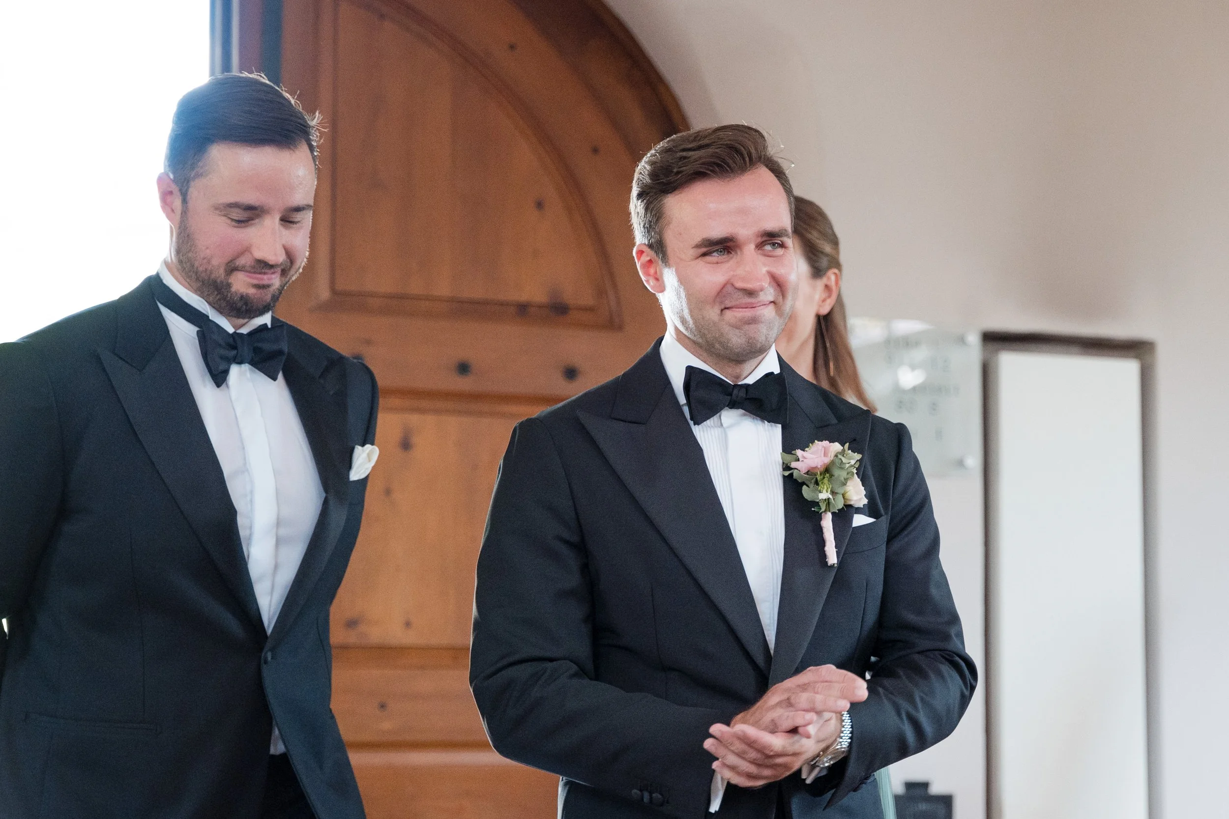 Zwei Männer in Anzügen mit Fliegen, einer trägt ein Kleid mit Blumen, die bei einer Hochzeit stehen, beide sind emotional. Eine Frau im Hintergrund. Innenraum mit Holzdekor.