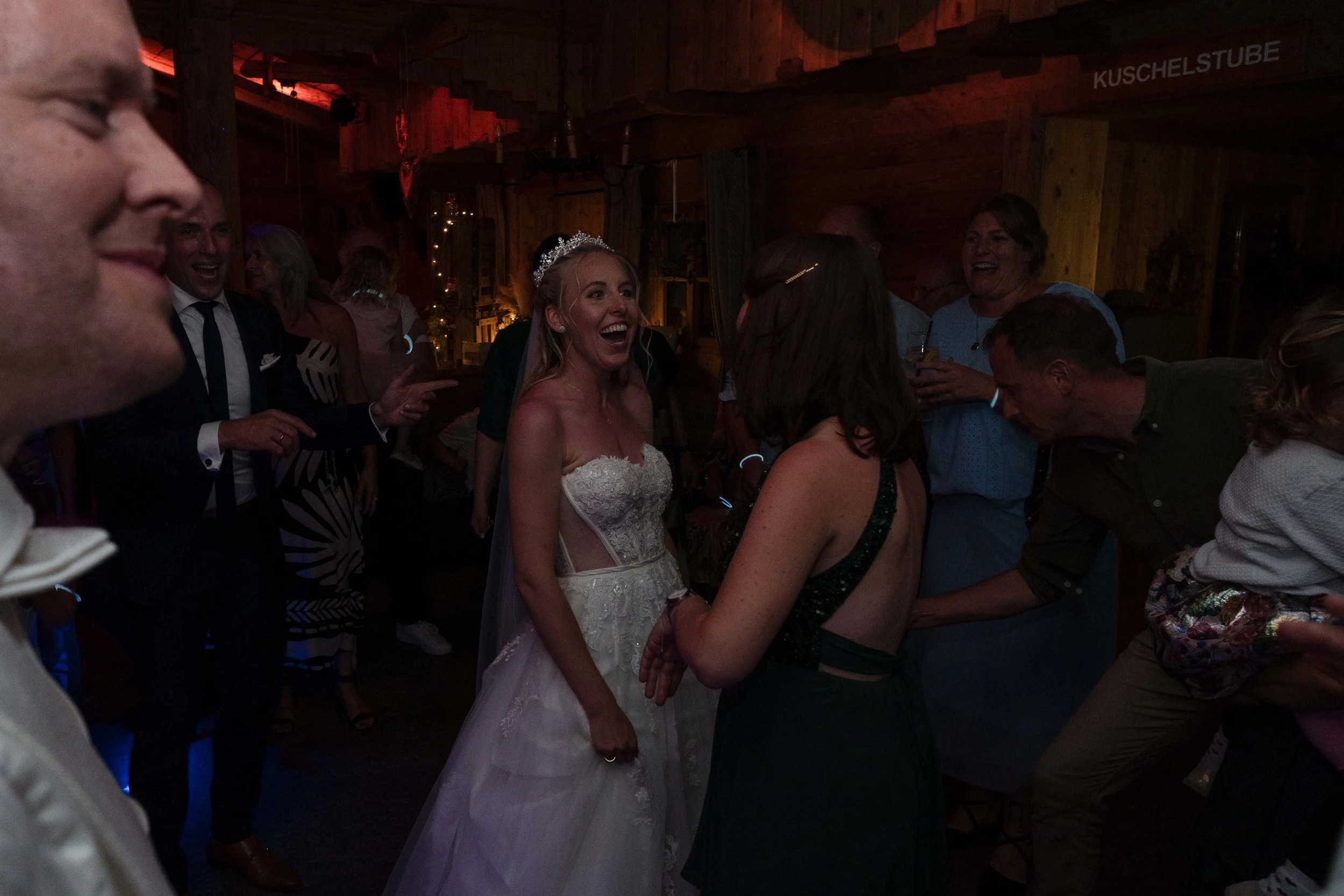 Menschen feiern in einer rustikalen Holzhütte. Im Mittelpunkt eine lachende Frau in Brautkleid mit Diadem, umgeben von weiteren Gästen.