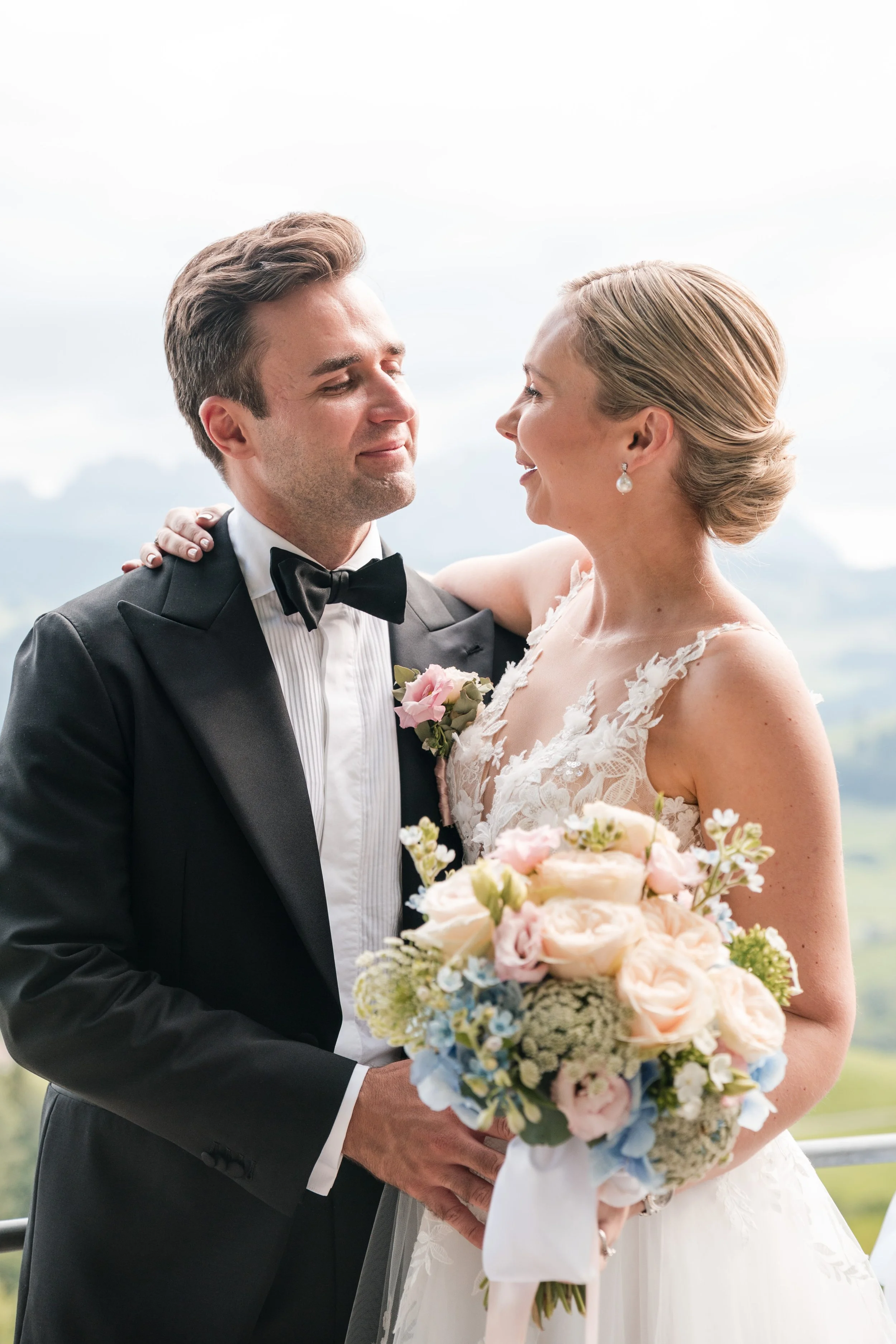 Ein Brautpaar bei einer Hochzeit, der Bräutigam trägt einen schwarzen Anzug und Schleife, die Braut in einem weißen Kleid mit Blumen, sie hält einen Blumenstrauß, im Hintergrund ist eine ländliche Landschaft sichtbar.