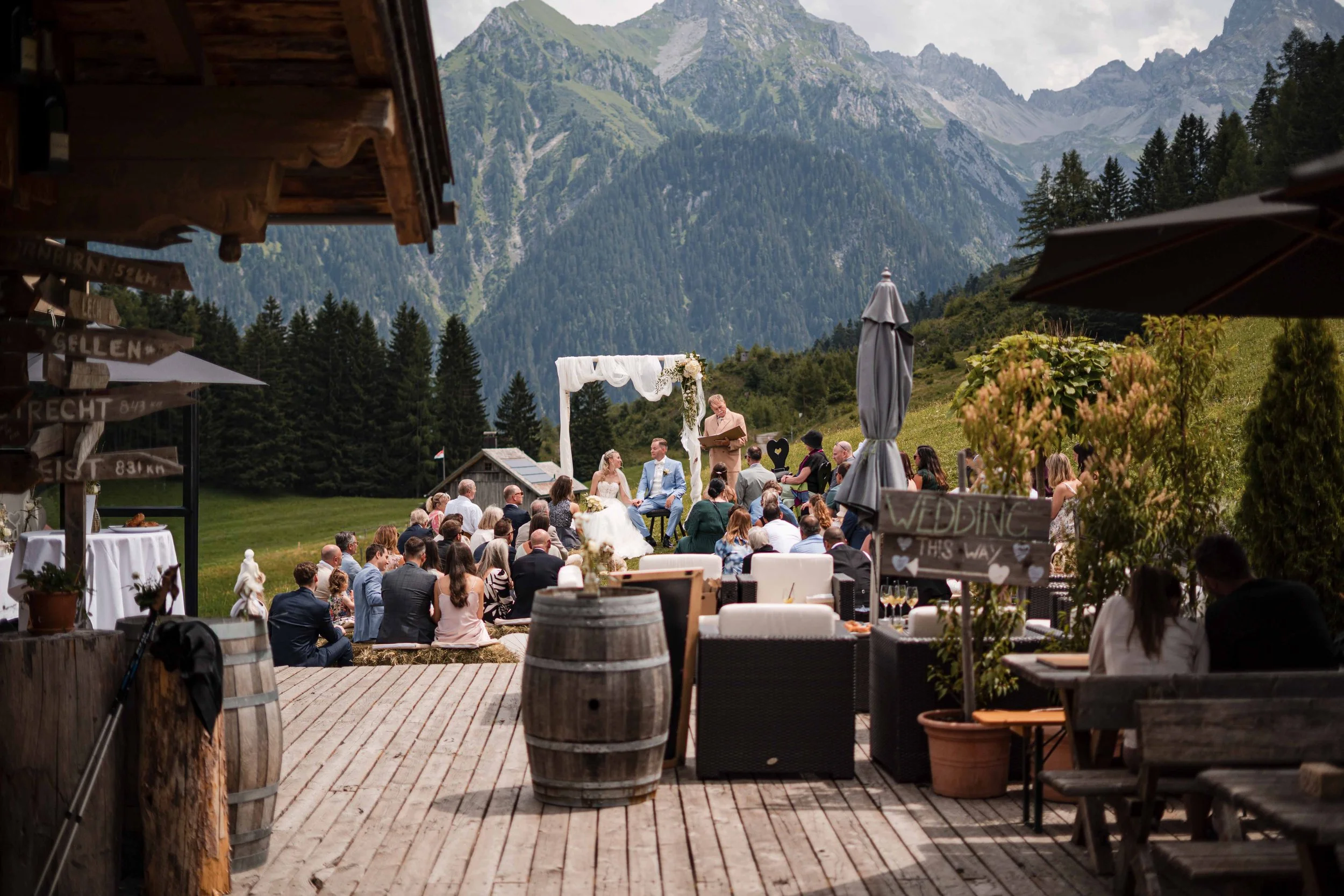Eine Hochzeit im Freien vor einer Bergkulisse mit vielen Gästen, einem Hochzeitspaar und einem Trauzeugen bei einer Zeremonie unter einem weißen Bogen. Im Vordergrund befinden sich Tische und Stühle, sowie Dekorationen und eine Tafel mit dem Schriftz