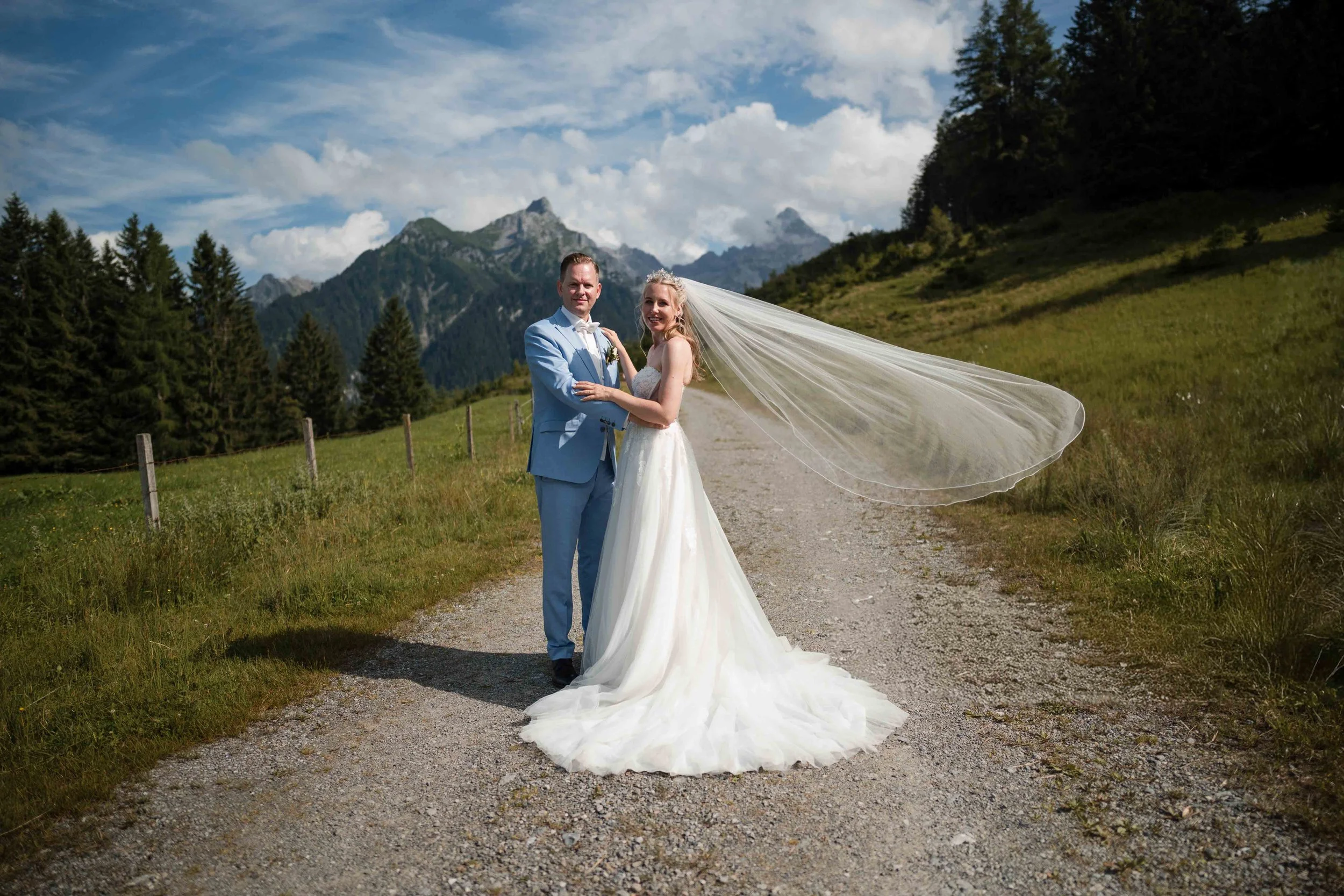 Ein Brautpaar in Hochzeitskleidung auf einem ländlichen Weg mit Bergen im Hintergrund, die Braut trägt ein langes weißes Kleid mit Schleier, der vom Wind bewegt wird, der Bräutigam trägt einen hellblauen Anzug.