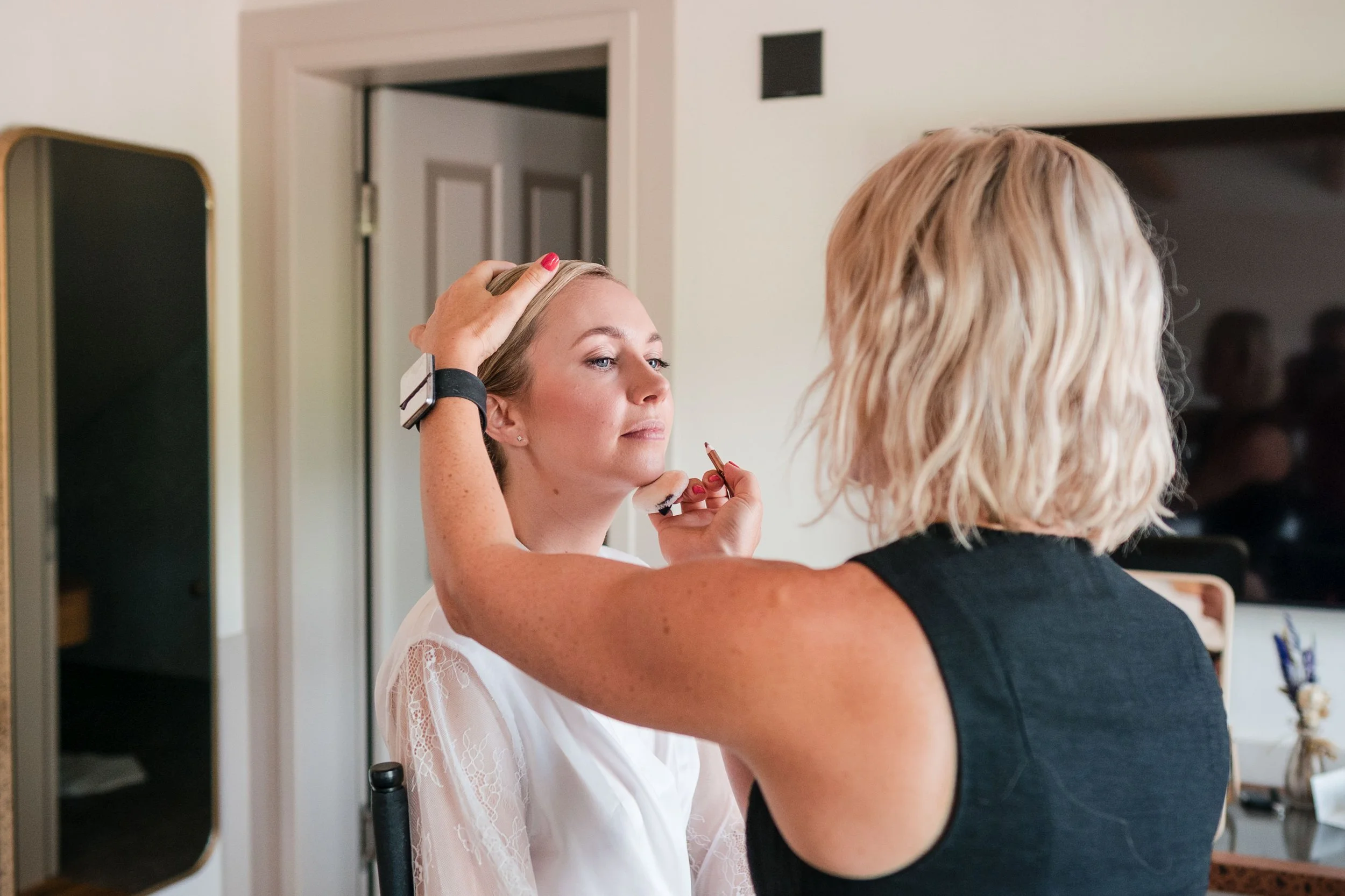 Eine Frau wird von einer anderen Frau geschminkt. Die geschminkte Frau sitzt auf einem Stuhl, während die andere Frau Pinsel und Make-up-Produkte verwendet.