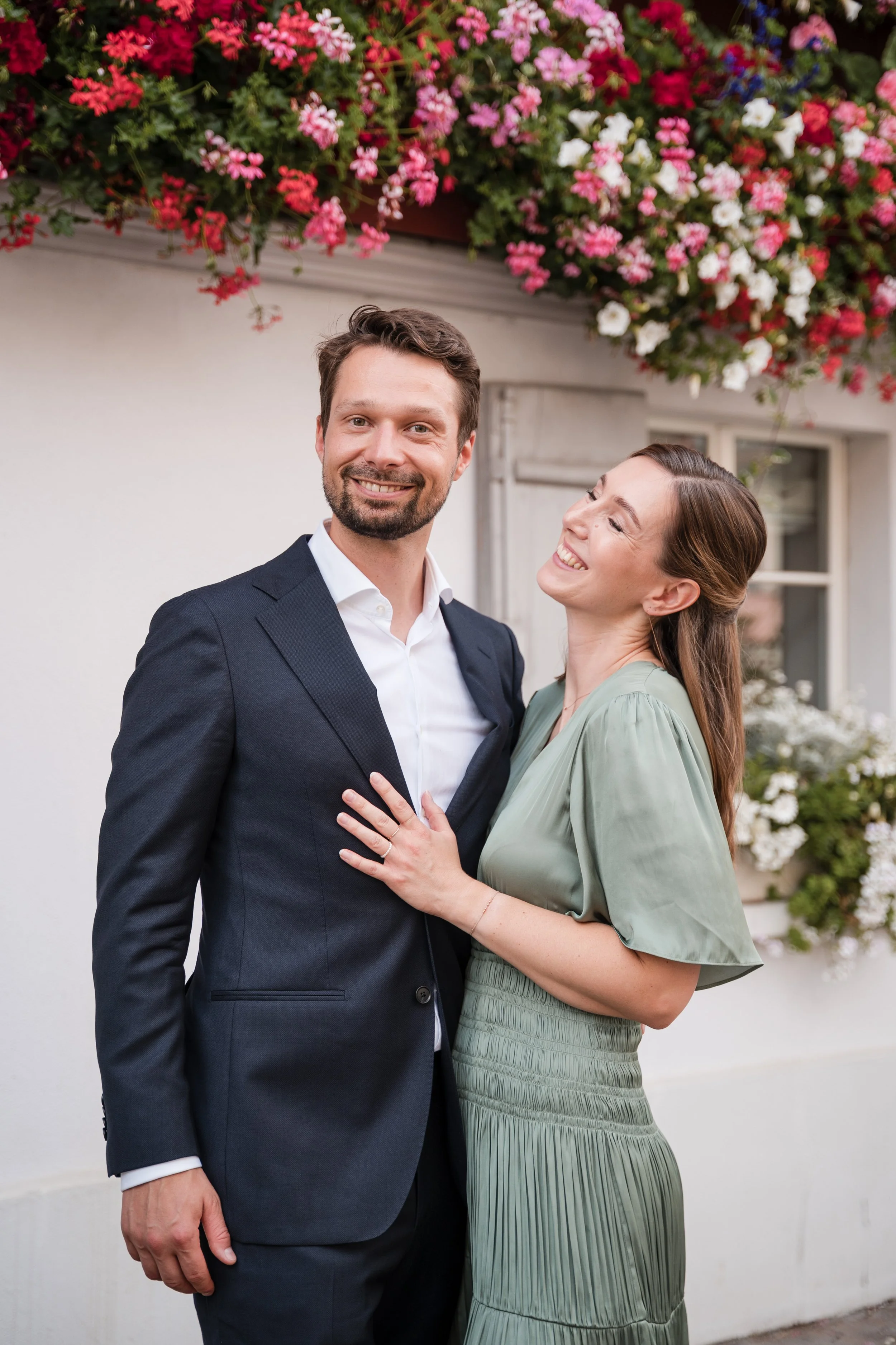 Ein lachendes Paar steht vor einer weißen Hauswand mit bunten Blumen im Hintergrund.