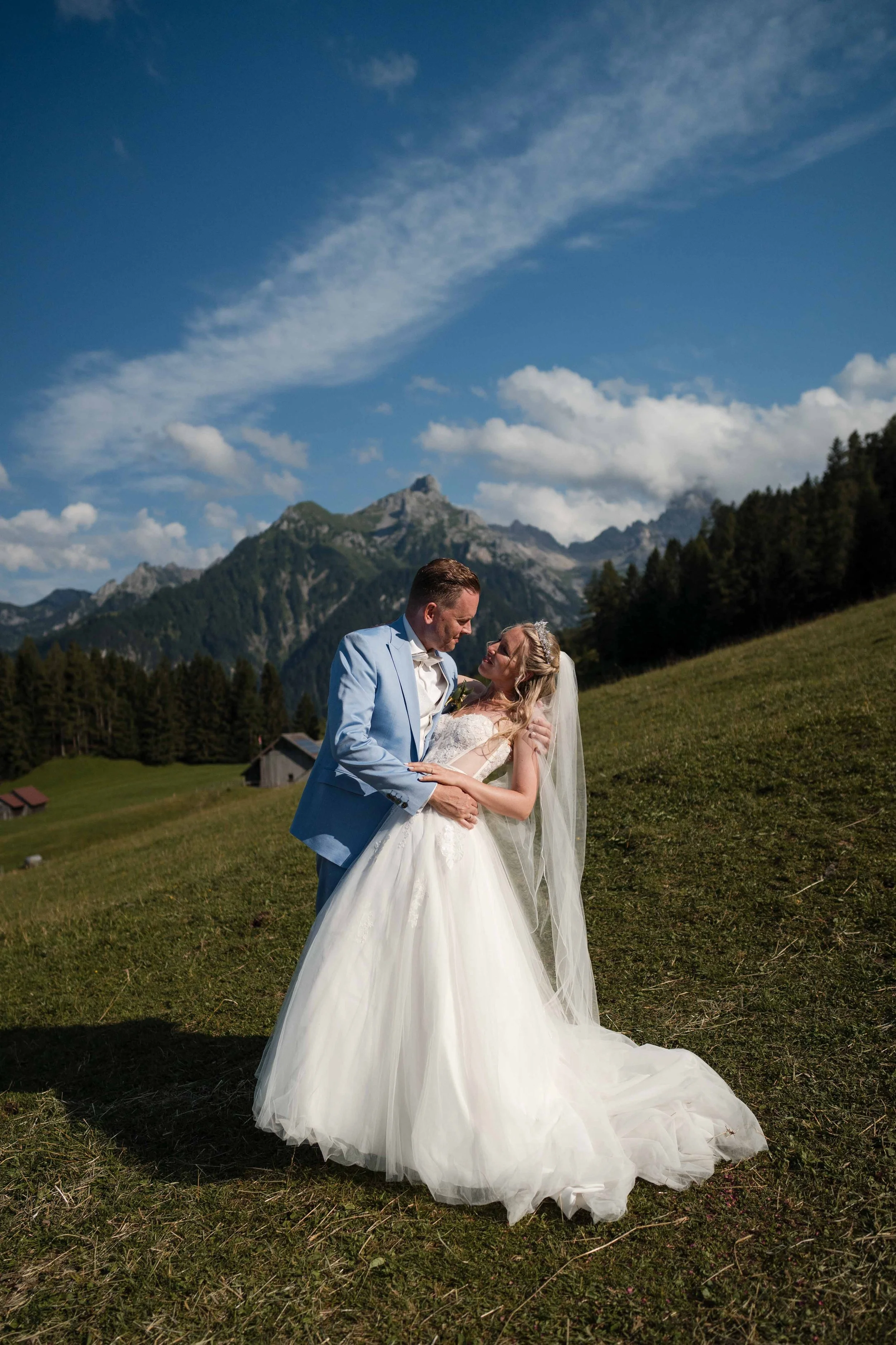 Ein Brautpaar in Hochzeitskleidung, das sich in einer malerischen, bergigen Landschaft umarmt, mit blauen Himmel und Wolken im Hintergrund.