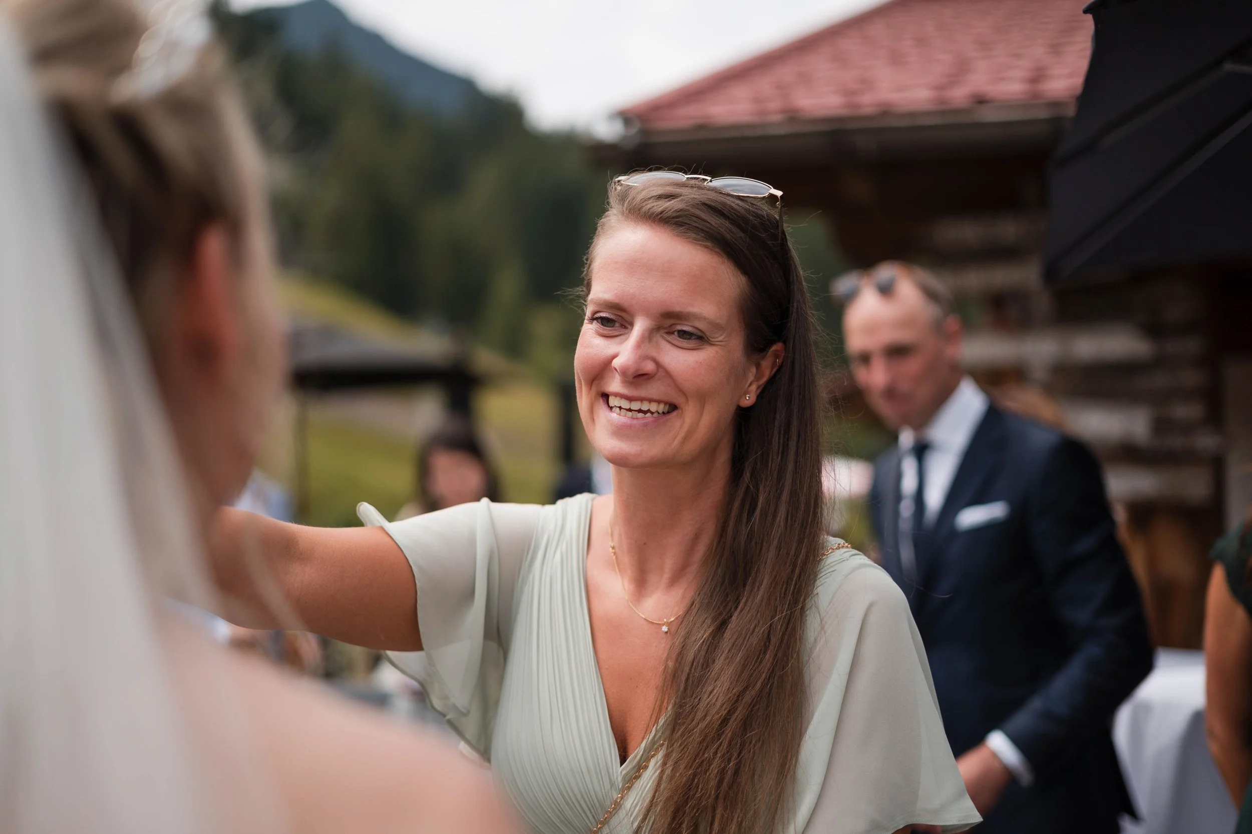 Eine lachende Frau beim Hochzeitsempfang, umgeben von Gästen im Freien mit Bergen im Hintergrund.