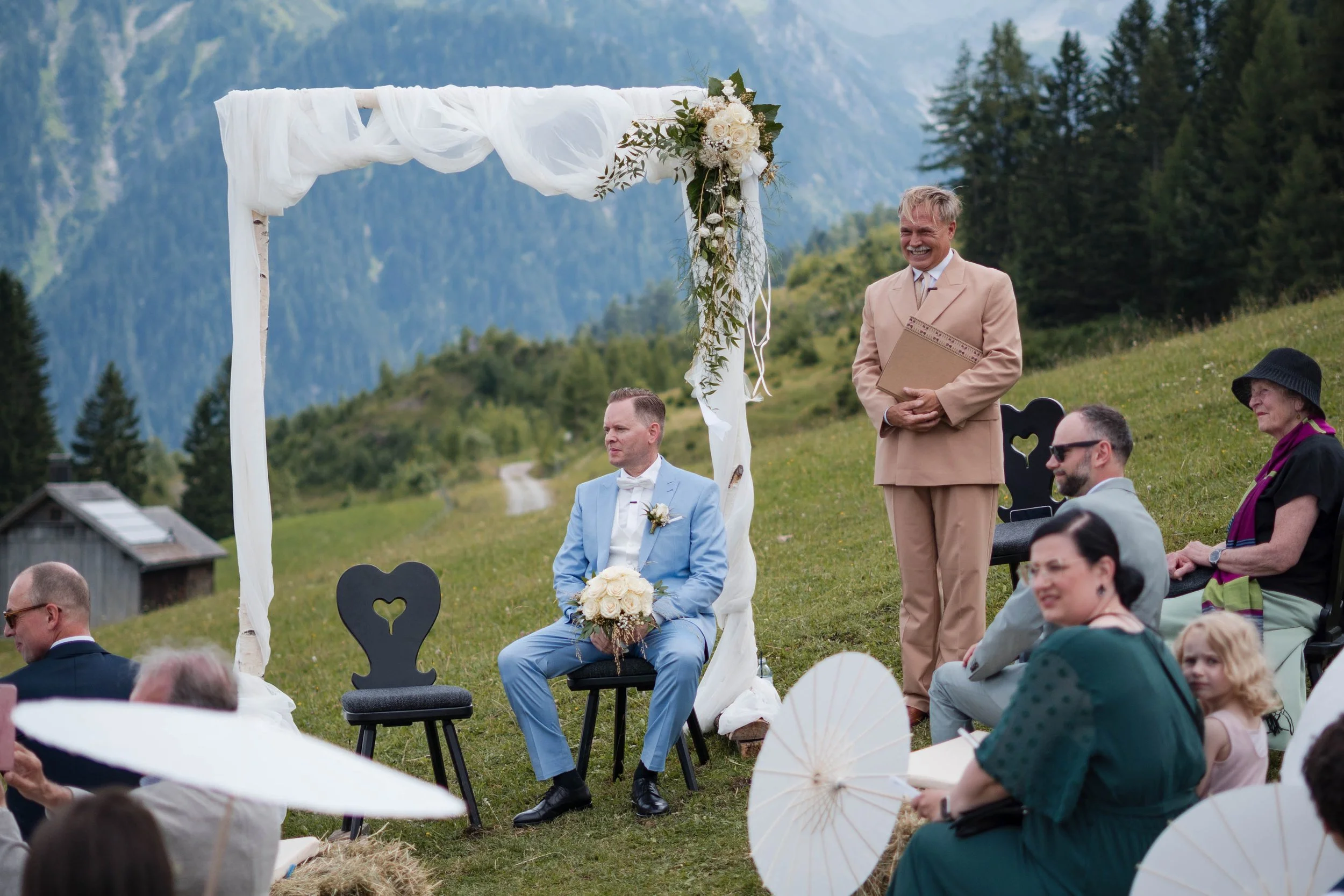 Hochzeit im Freien mit Bergen im Hintergrund, zeremonielle Dekoration mit weißem Tuch und Blumen, Braut und Bräutigam sitzen vor einer Gruppe von Gästen, einige Gäste halten Papier- oder Sonnenregenschirme, es ist sonniges Wetter.