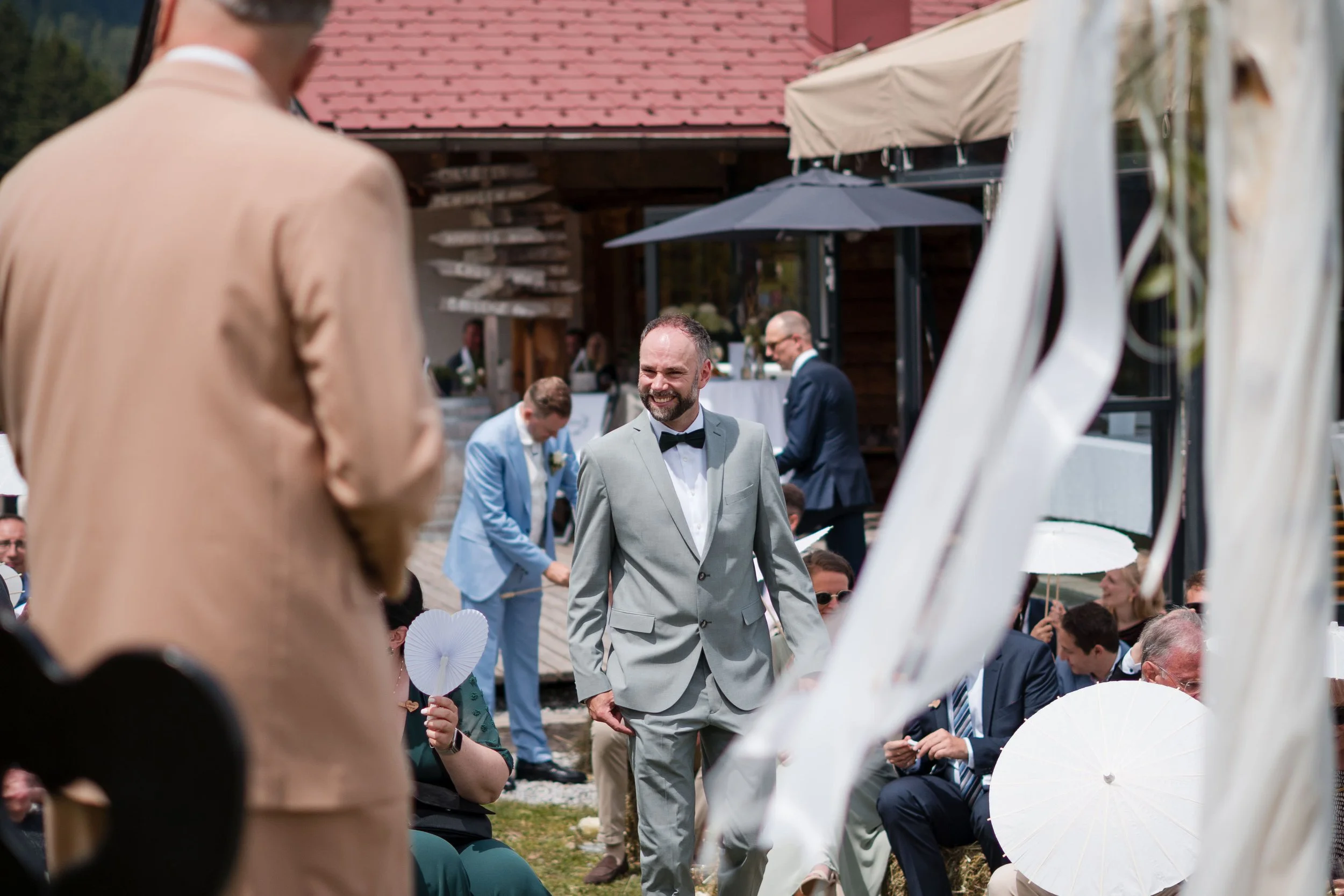 Ein lächelnder Mann in einem hellgrauen Anzug und Fliegenknoten bei einer Hochzeit im Freien, umgeben von Gästen mit Regenschirmen.