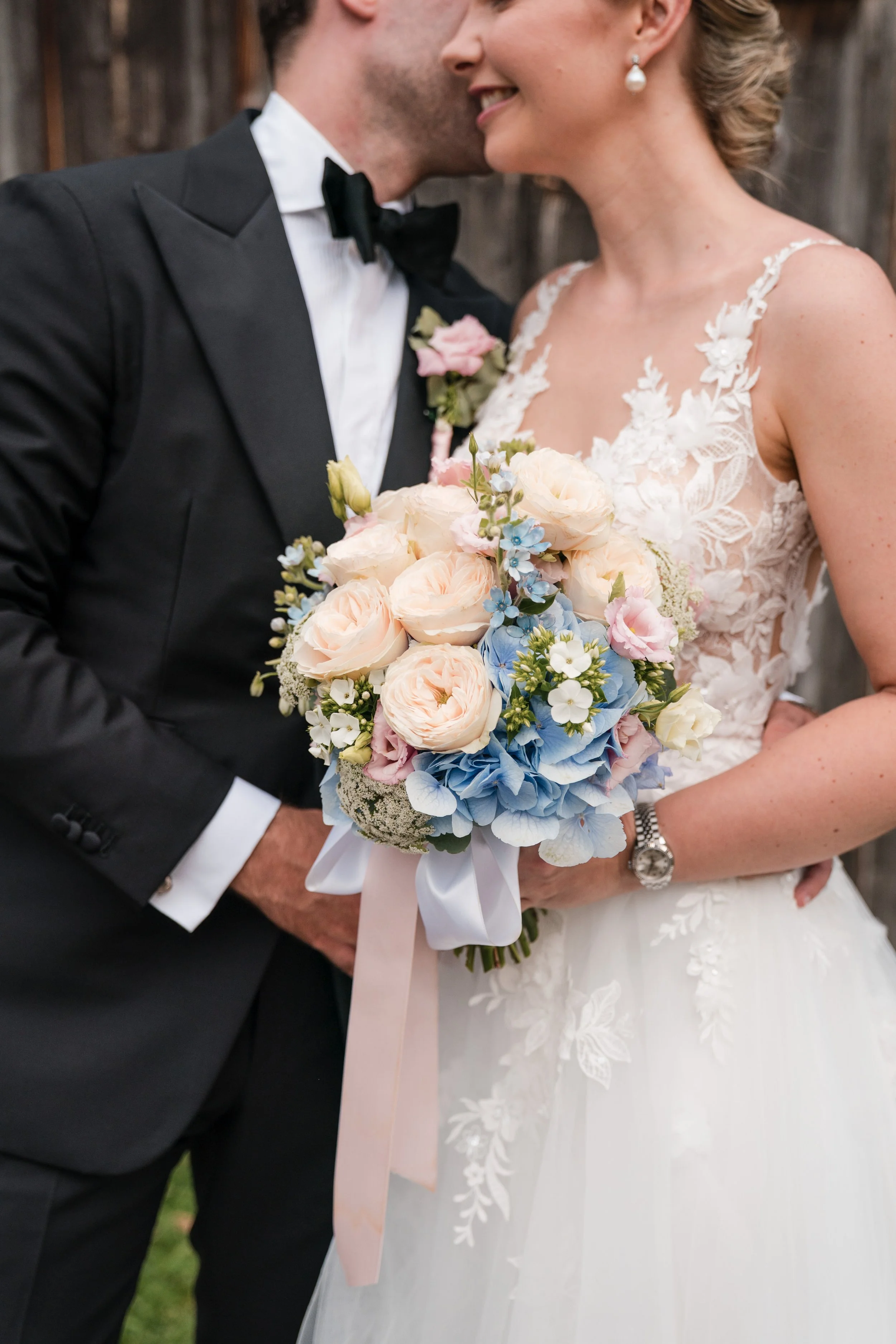 Braut und Bräutigam bei ihrer Hochzeit, umarmt und lächelnd, Braut hält einen Blumenstrauß mit blauen, weißen und rosa Blumen.