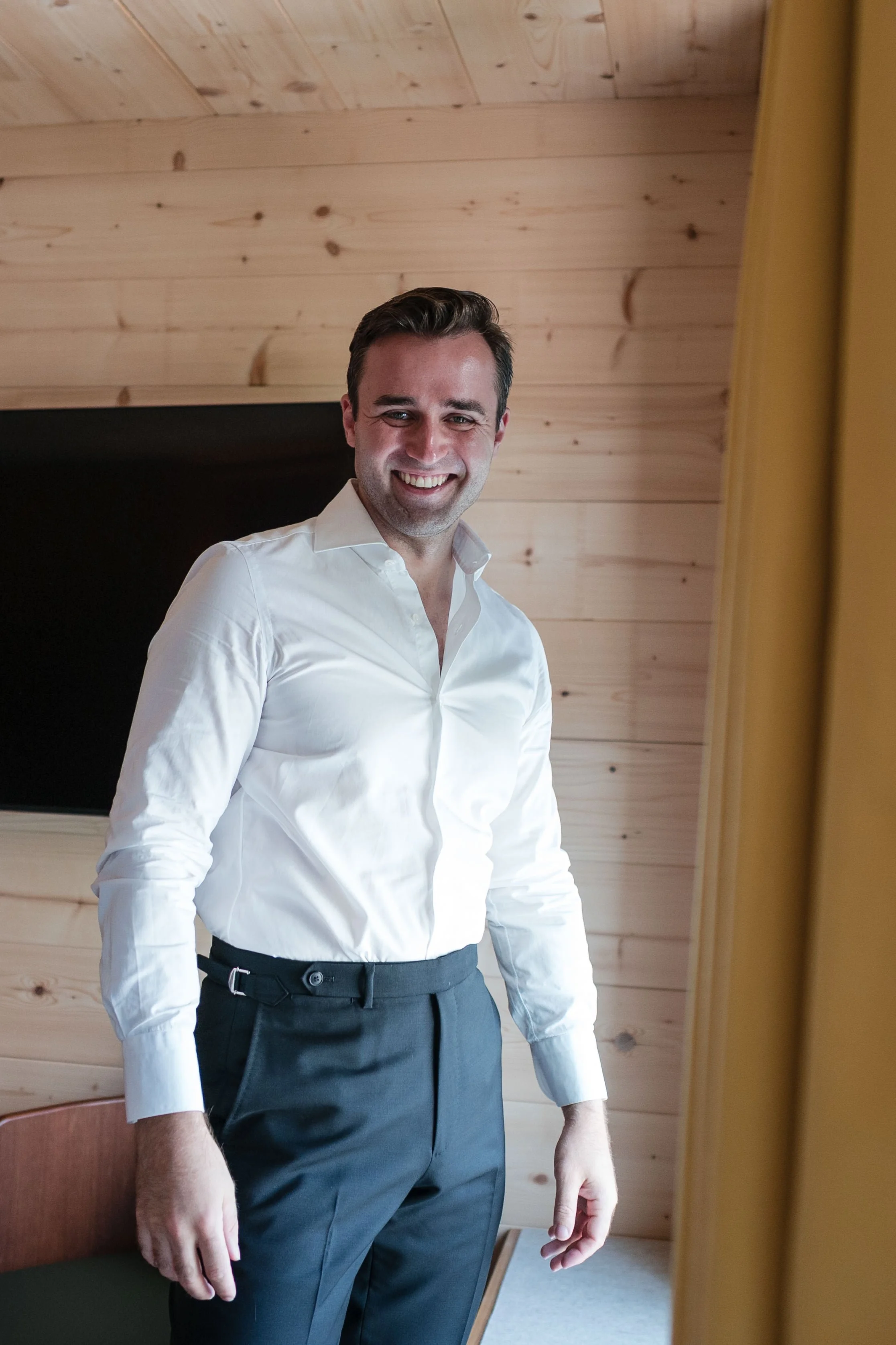 Ein lachender Mann in einem weißen Hemd und dunklen Hosen steht in einem Raum mit Holzverkleidung an den Wänden.