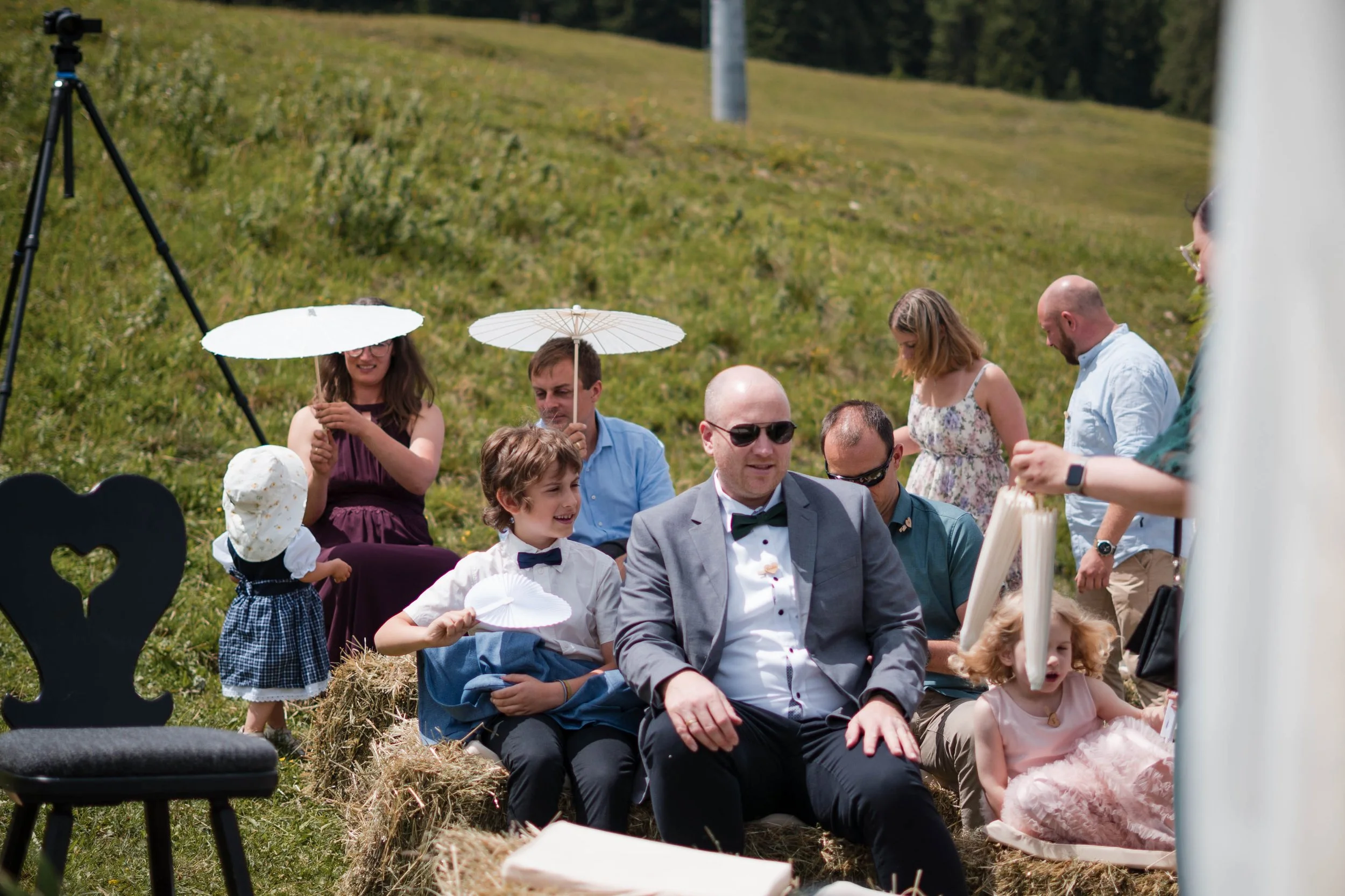 Gruppe von Menschen, die im Freien sitzen, teils mit Sonnenschirmen, bei einer Veranstaltung auf einer Wiese.