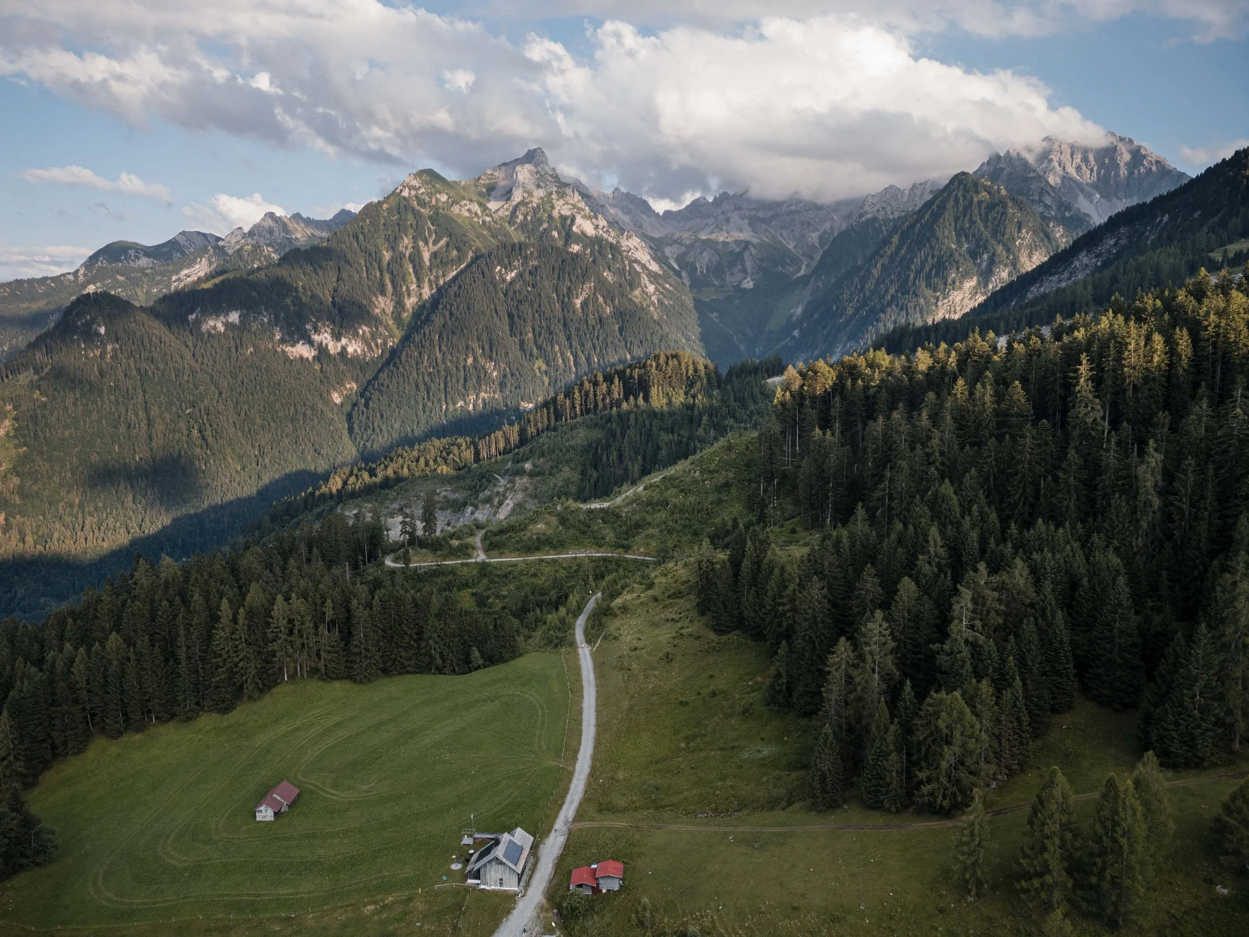 Eine grüne Hügellandschaft mit einer kleinen Farm und einer kurvenreichen Straße, umgeben von einem dichten Wald und Bergen im Hintergrund unter einem bewölkten Himmel.