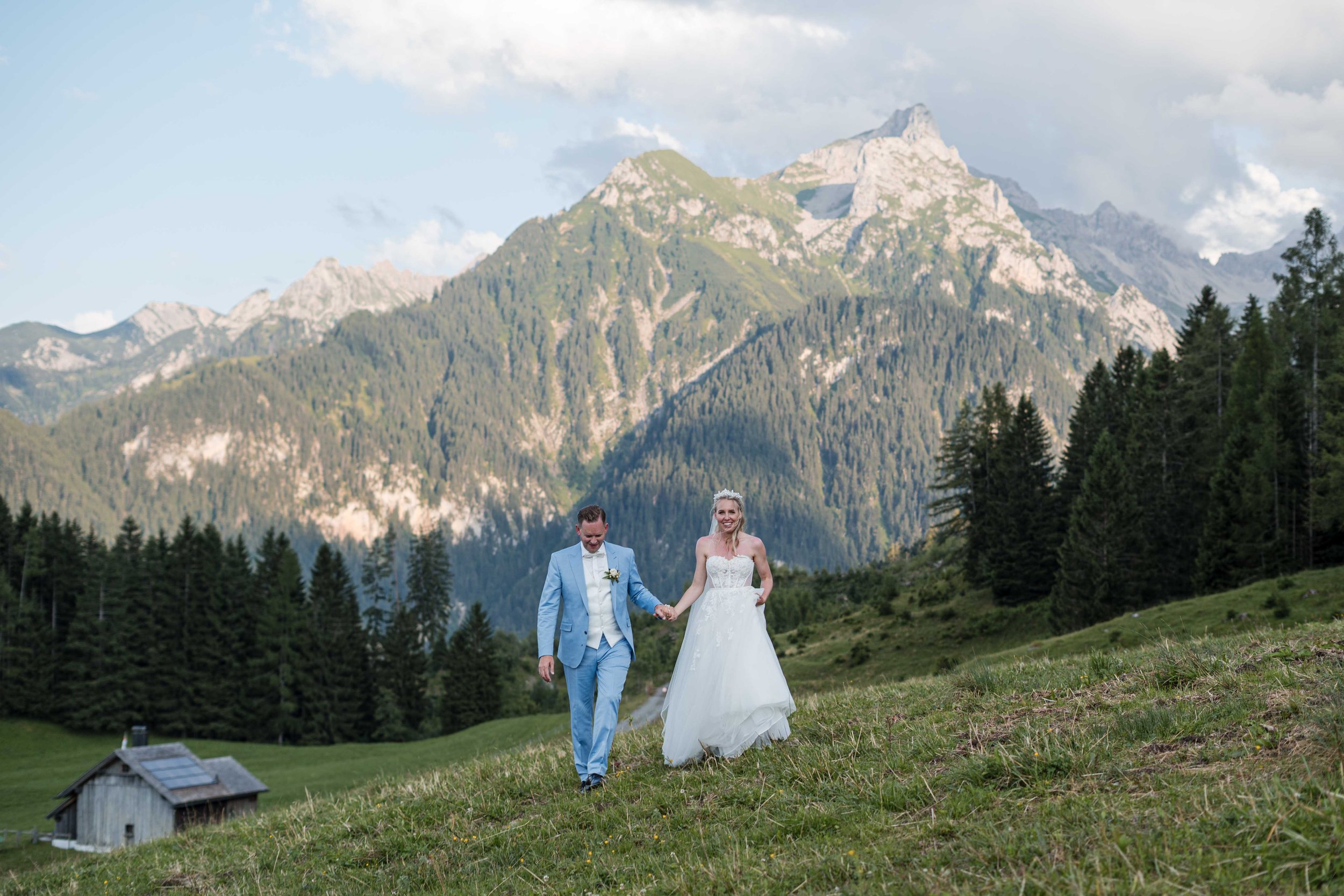 Ein Brautpaar hält Händchen beim Spazierengehen in einer grünen Berglandschaft mit hohen Bergen und Tannen im Hintergrund.