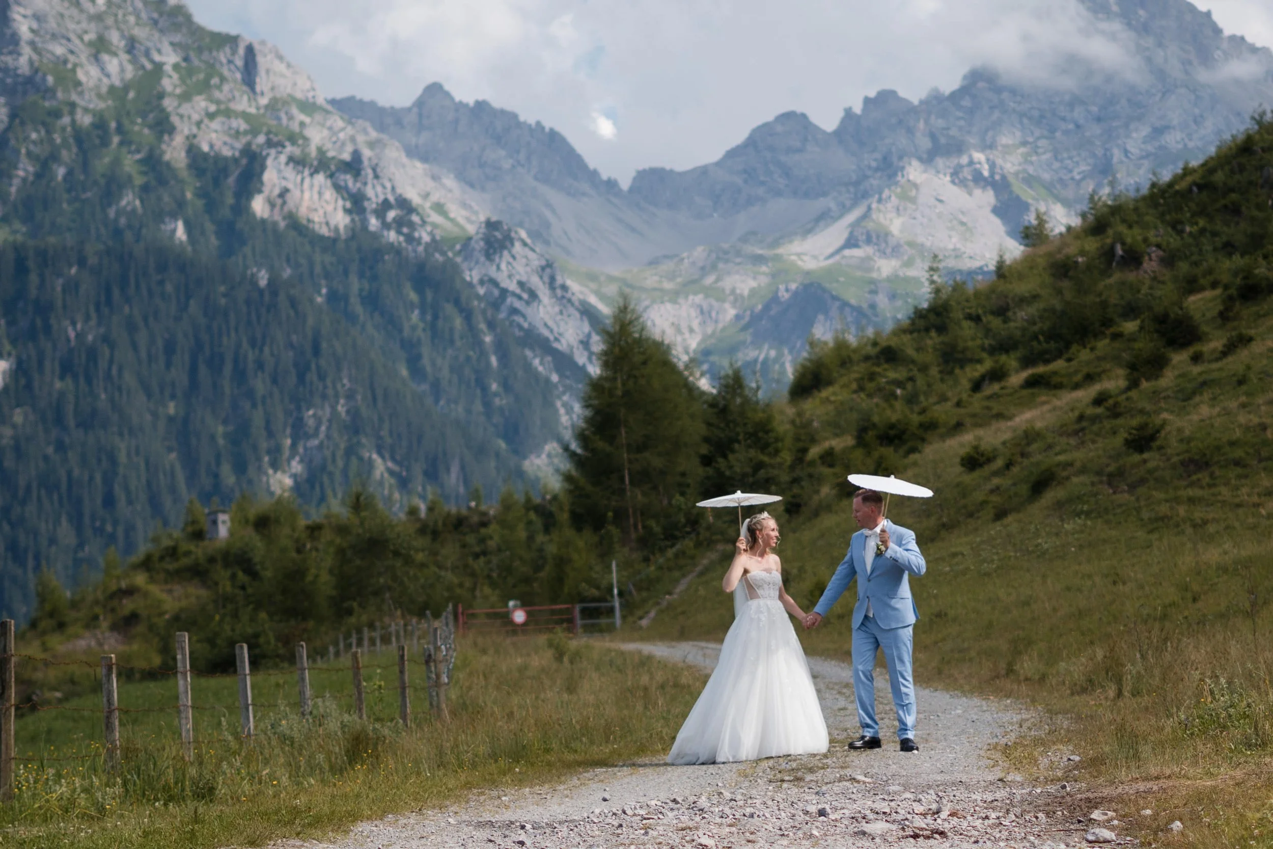 Ein Brautpaar hält Händchen und trägt weiße und blaue Anzüge unter weißen Sonnen-Schirmen in einer malerischen Berglandschaft.