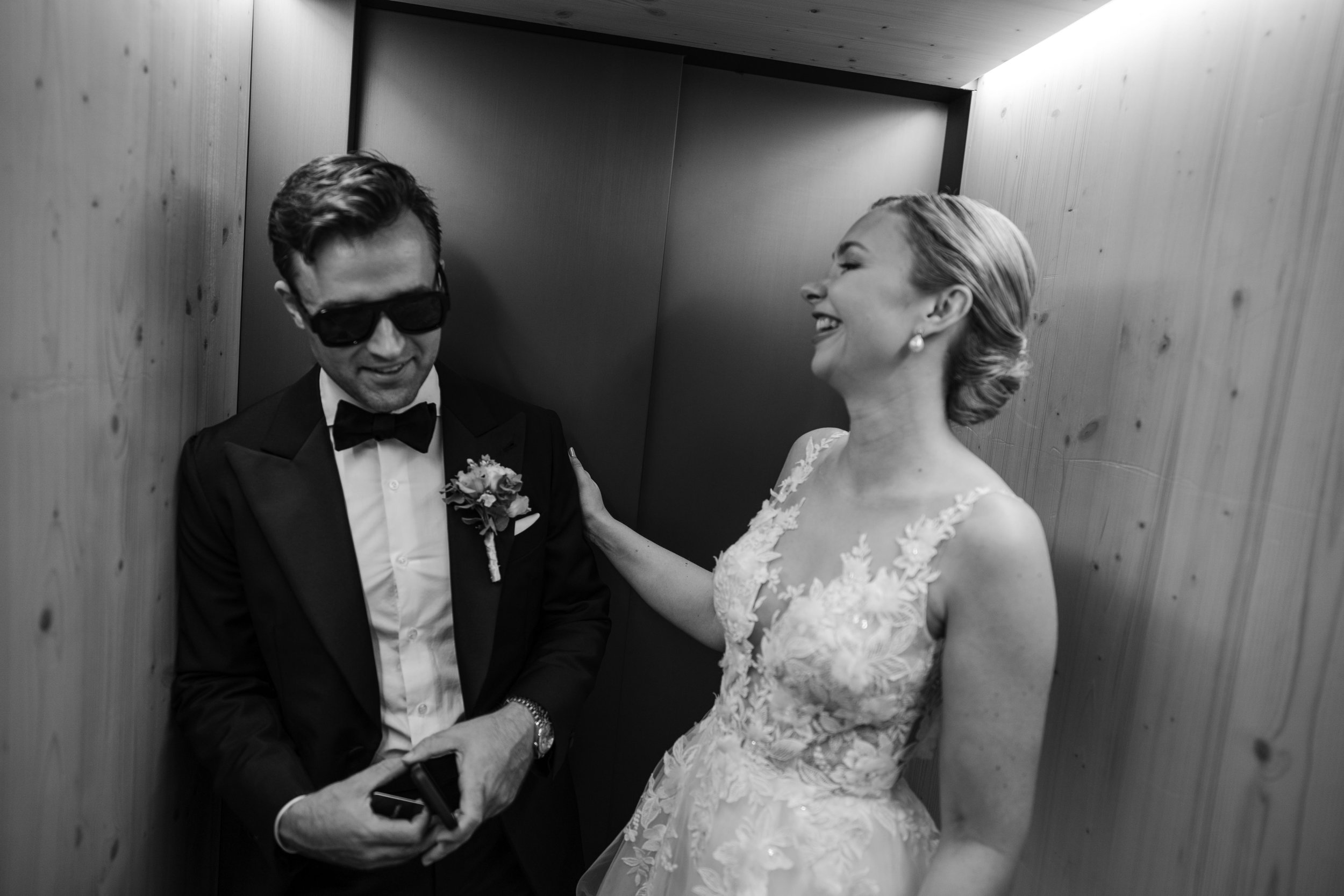 Ein Mann im Smoking und eine Frau in einem Hochzeitskleid lachen und unterhalten sich in einem Aufzug aus Holz.