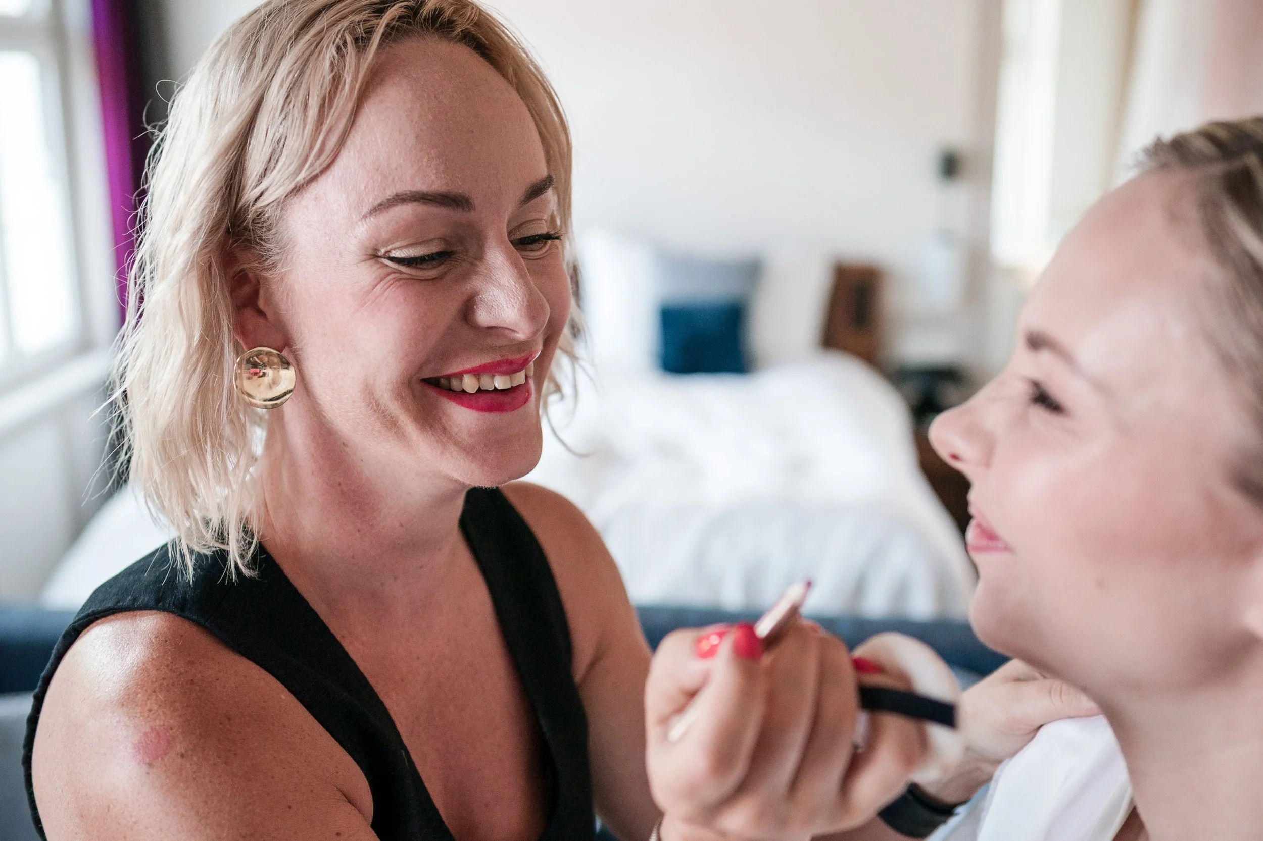 Eine Frau benutzt einen Pinsel, um Make-up auf eine andere Frau aufzutragen, beide lachen und sind im Schlafzimmer.