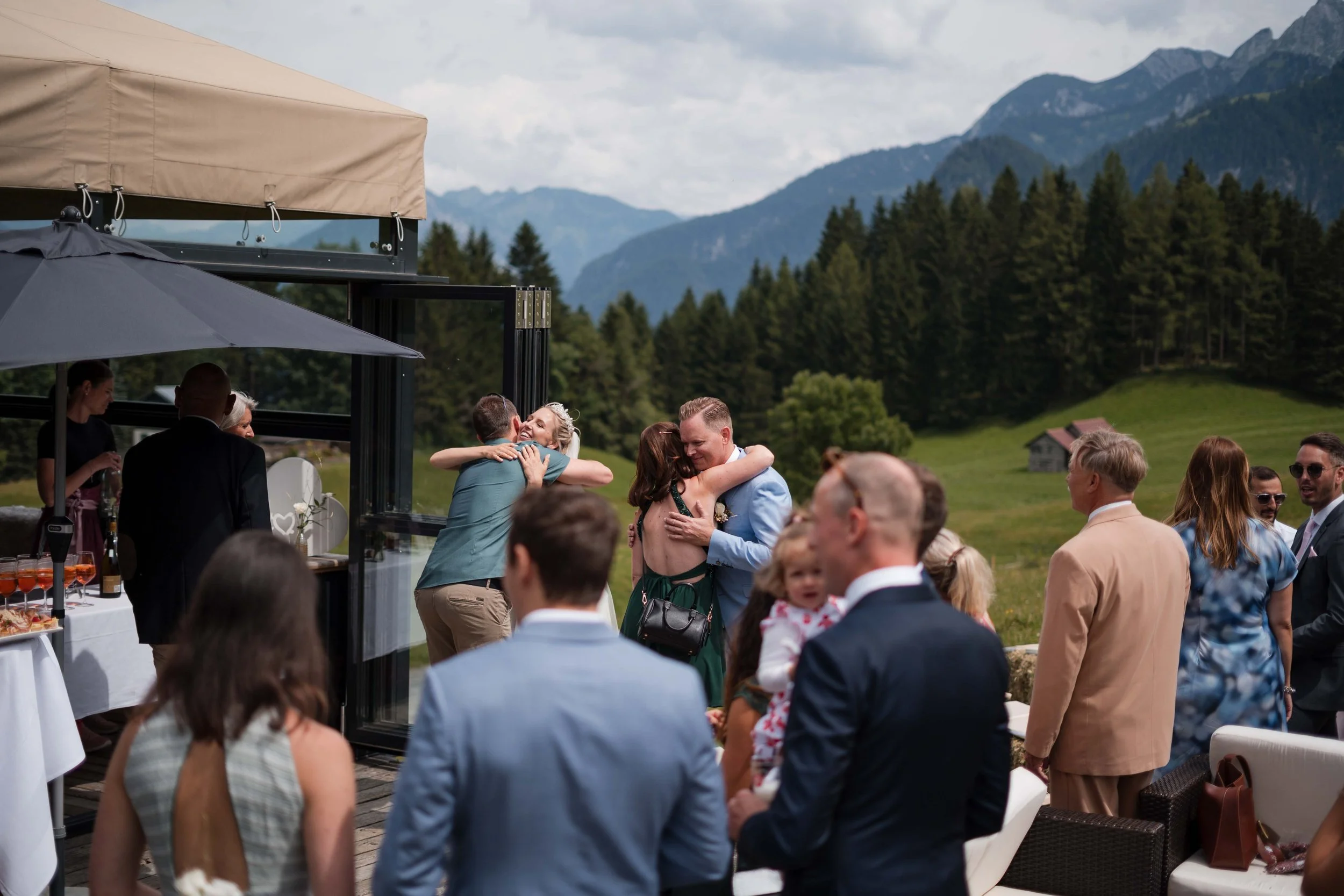 Hochzeit im Freien mit Gästen, die sich umarmen und feiern, vor einer bergigen Landschaft mit grünen Wiesen und Bäumen.