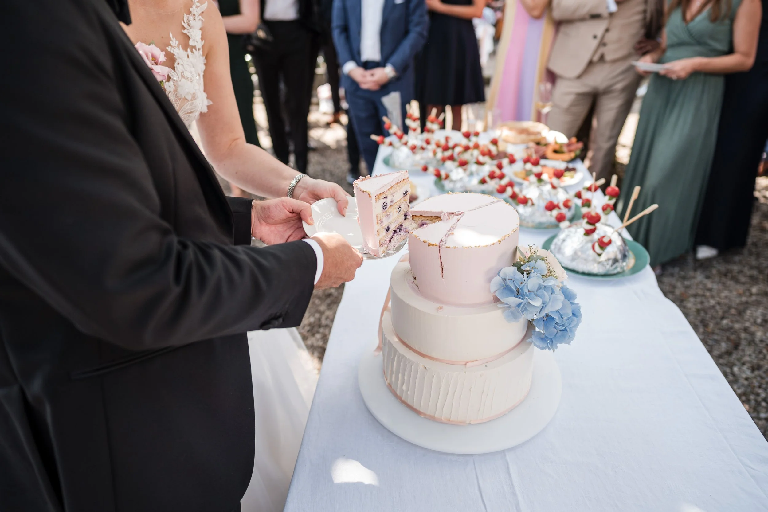 Hochzeitstorte mit mehreren Schichten, rosa und weiß, mit blauer Hortensie, wird bei einer Hochzeit angeschnitten, umgeben von Gästen.