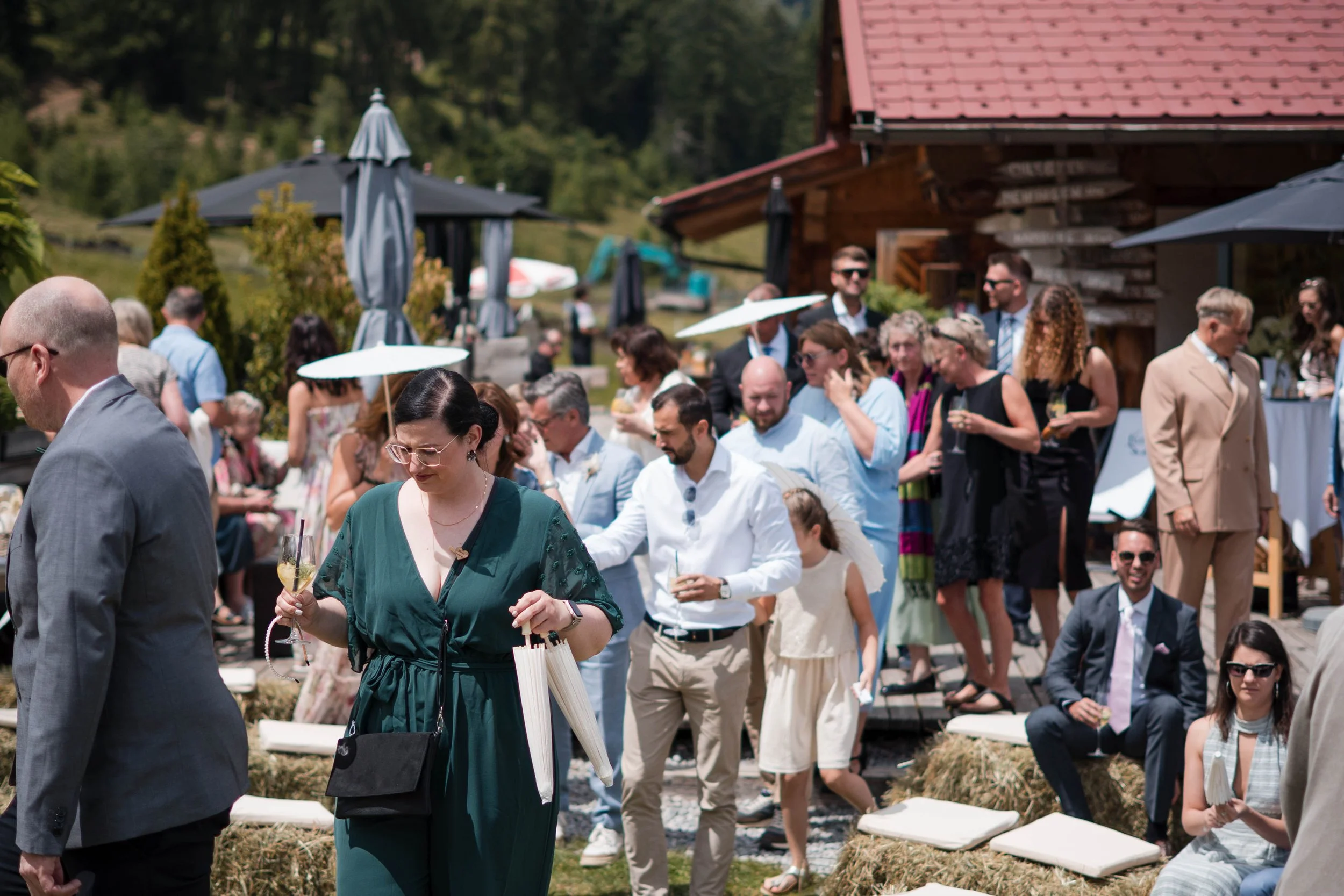 Menschen bei einer Outdoor-Hochzeit, einige mit Regenschirmen, im sonnigen Garten auf Strohballen, im Hintergrund Bäume und ein Holzhaus.