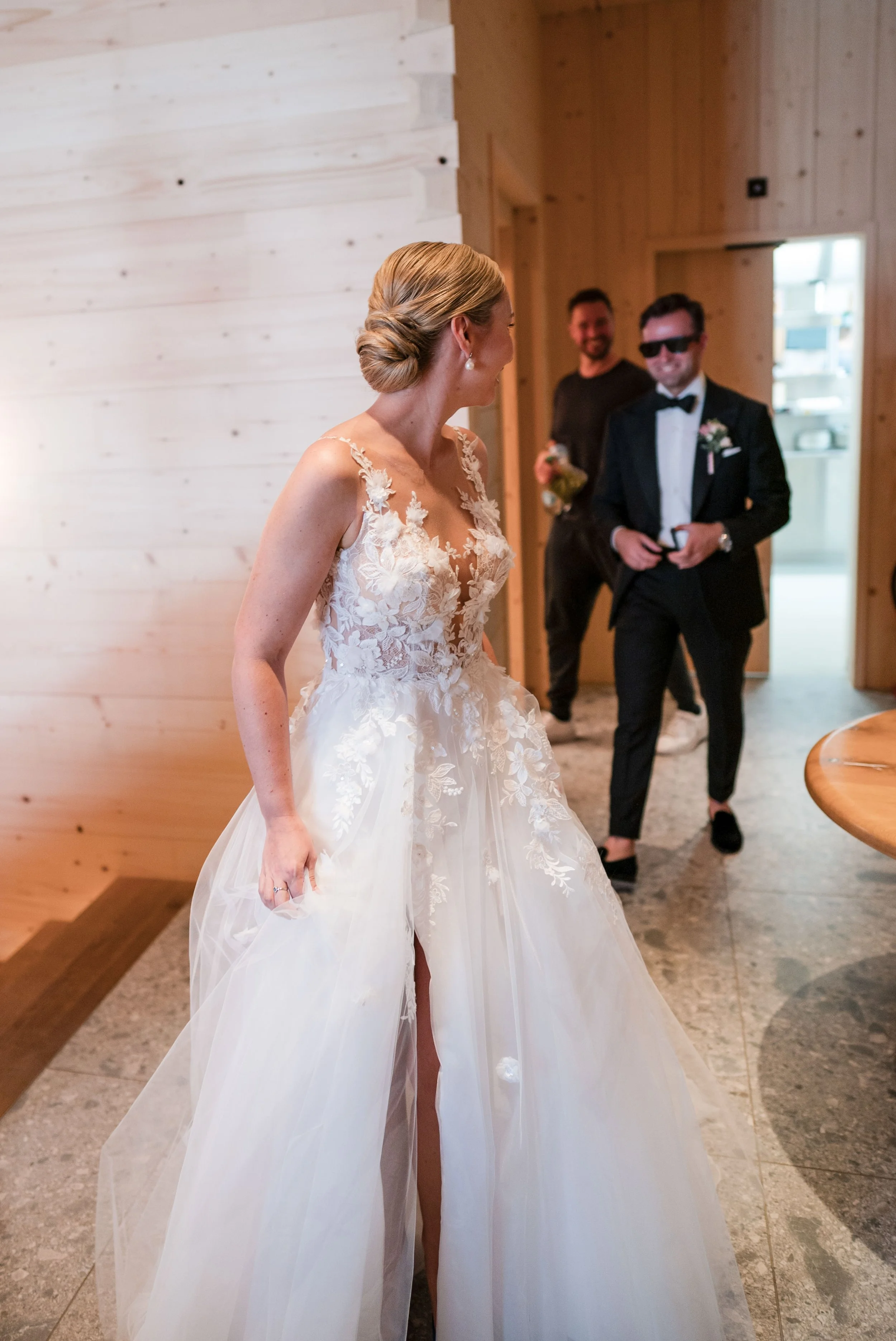 Braut in einem weißen, eleganten Hochzeitskleid mit floralem Muster und hohem Beinschlitz, lachend im Gespräch mit zwei Männern im Anzug im Holzhaus.