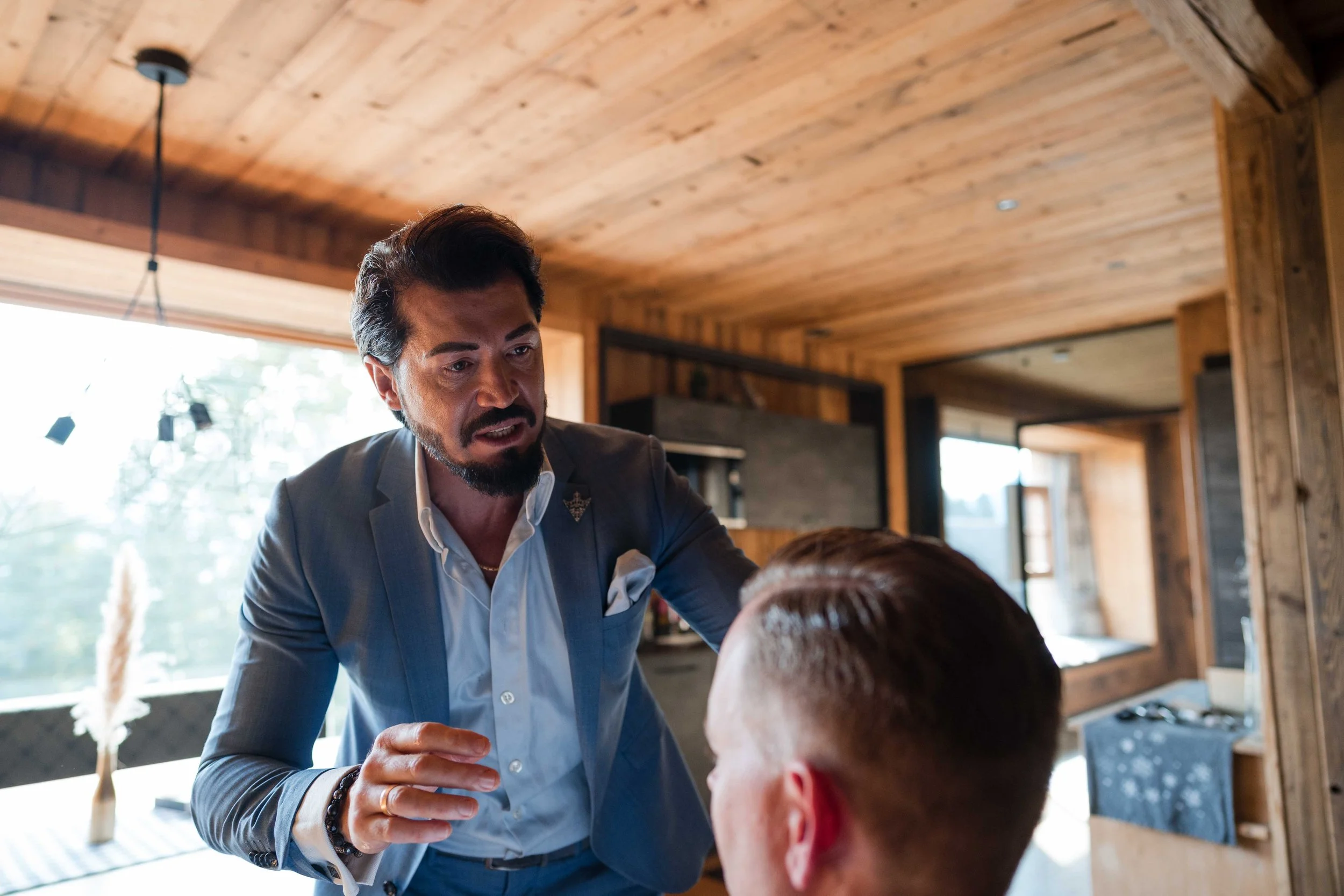 Ein Mann in einem grauen Anzug spricht mit einer Person mit kurzen Haaren in einer modernen, holzverkleideten Wohnung.