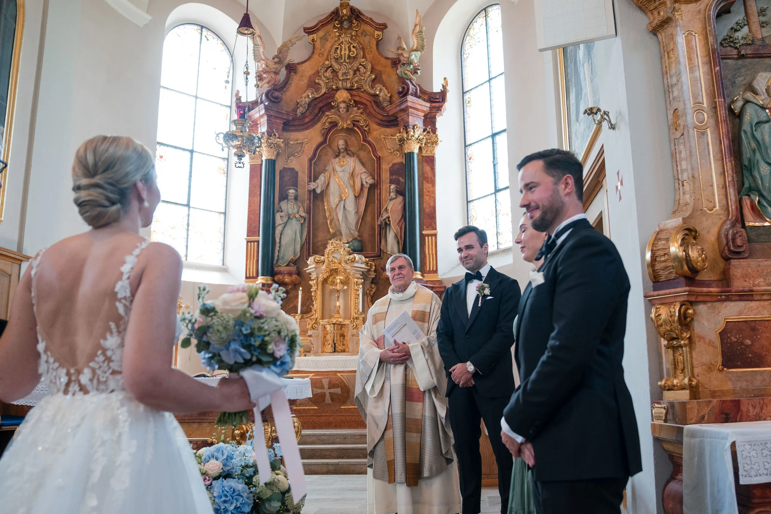 Ein Brautpaar bei einer Hochzeit in einer Kirche, mit einem Priester und zwei Männern, im Hintergrund ein barockes Altarbild mit goldener Verzierung.