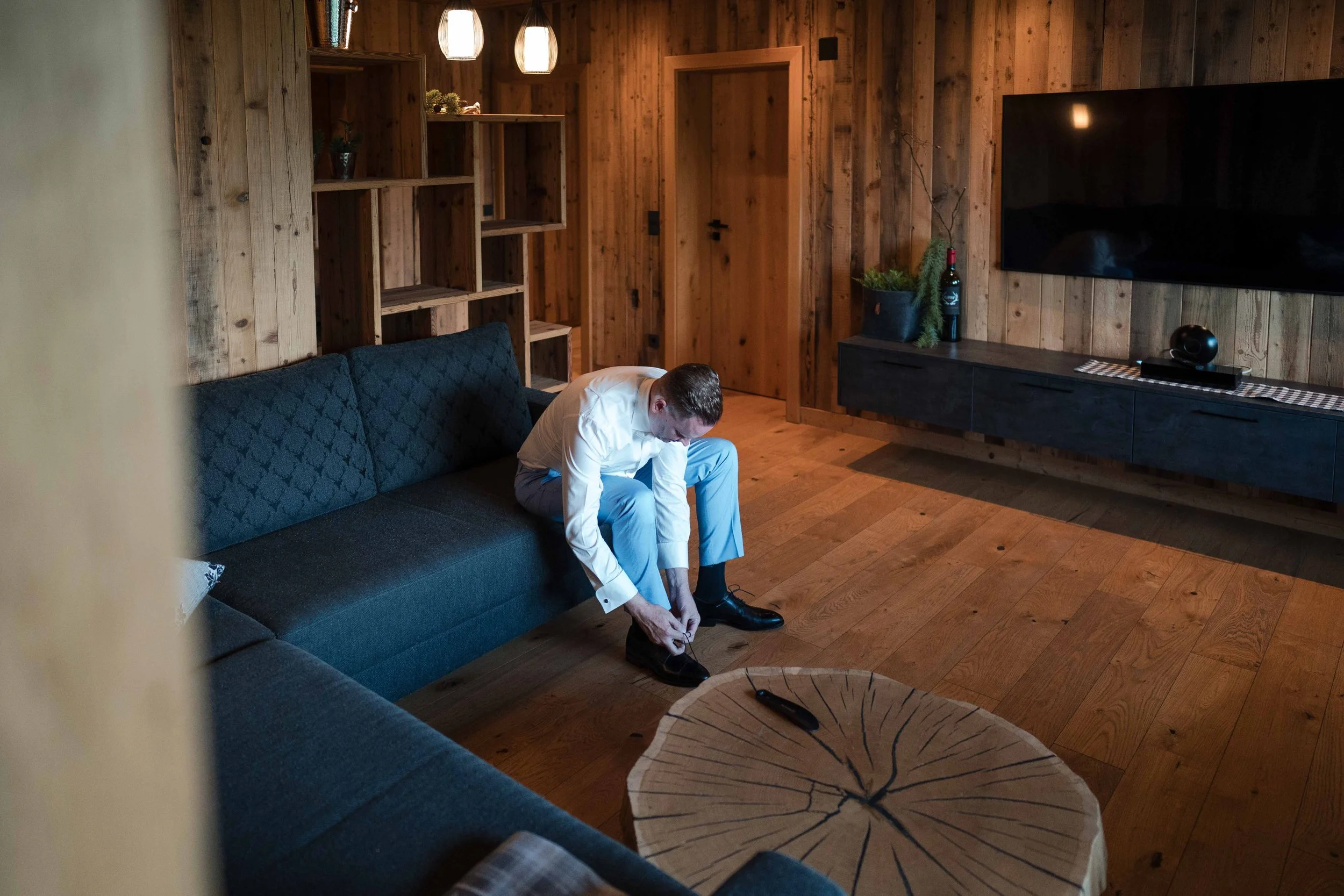Ein Mann im weißen Hemd und grauen Hosen sitzt auf einem dunklen Sofa und bindet seine schwarzen Schuhe im Wohnzimmer mit Holzwänden und Holzboden.