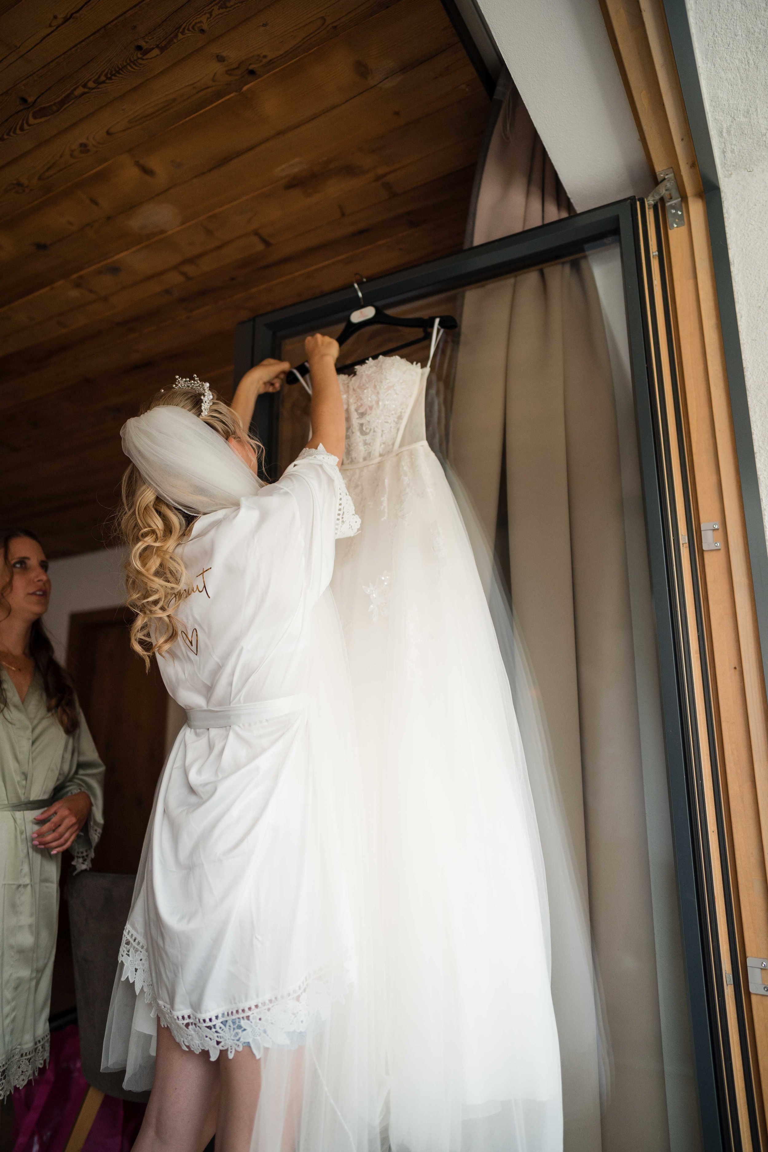 Frau in Brautkleid und Morgenmantel hängt ein Brautkleid auf einen Vorhanghaken. Eine andere Frau beobachtet sie.