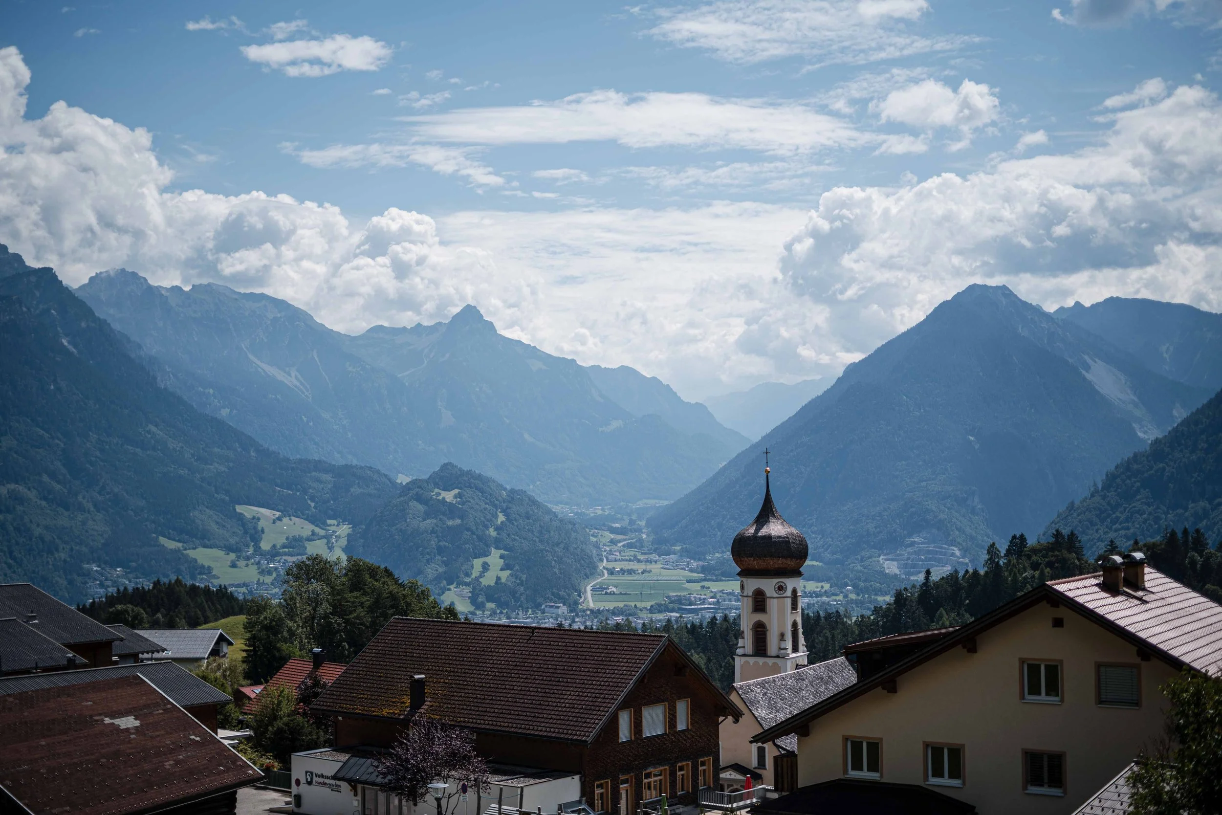 Blick auf ein Dorf in den Bergen mit einem Kirchturm vor einer beeindruckenden Berglandschaft und Wolken am Himmel.