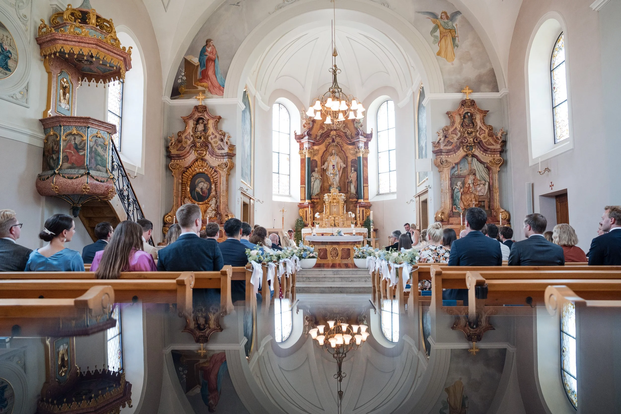 Innenansicht einer Kirche während einer Zeremonie, mit liturgischem Altar im Hintergrund, von Menschen in formeller Kleidung mit Blick auf das Altar. Die Kirche ist hell erleuchtet durch große Fenster, mit kunstvollen barocken Holzdekorationen und Wa