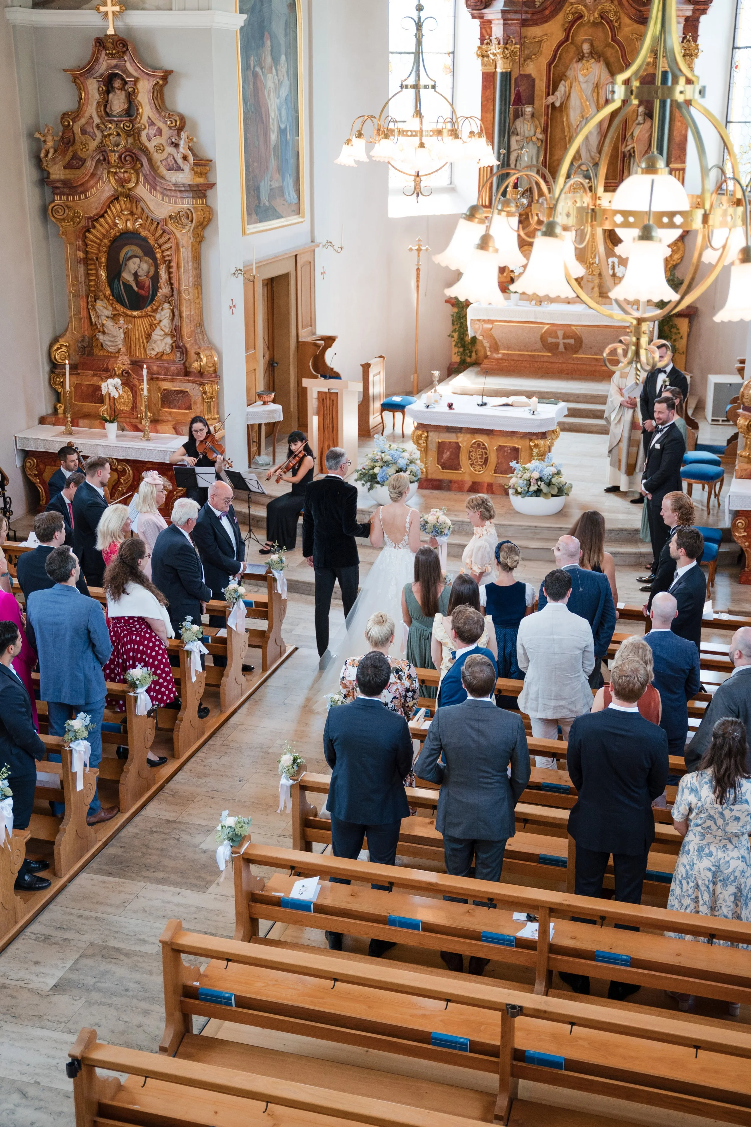 Ein Hochzeitsgottesdienst in einer kirchlichen Umgebung mit Brautpaar, Gästen und Musikern, die in einer kunstvoll dekorierten Kirche mit großen Chandelieren und religiösen Bildern stattfindet.