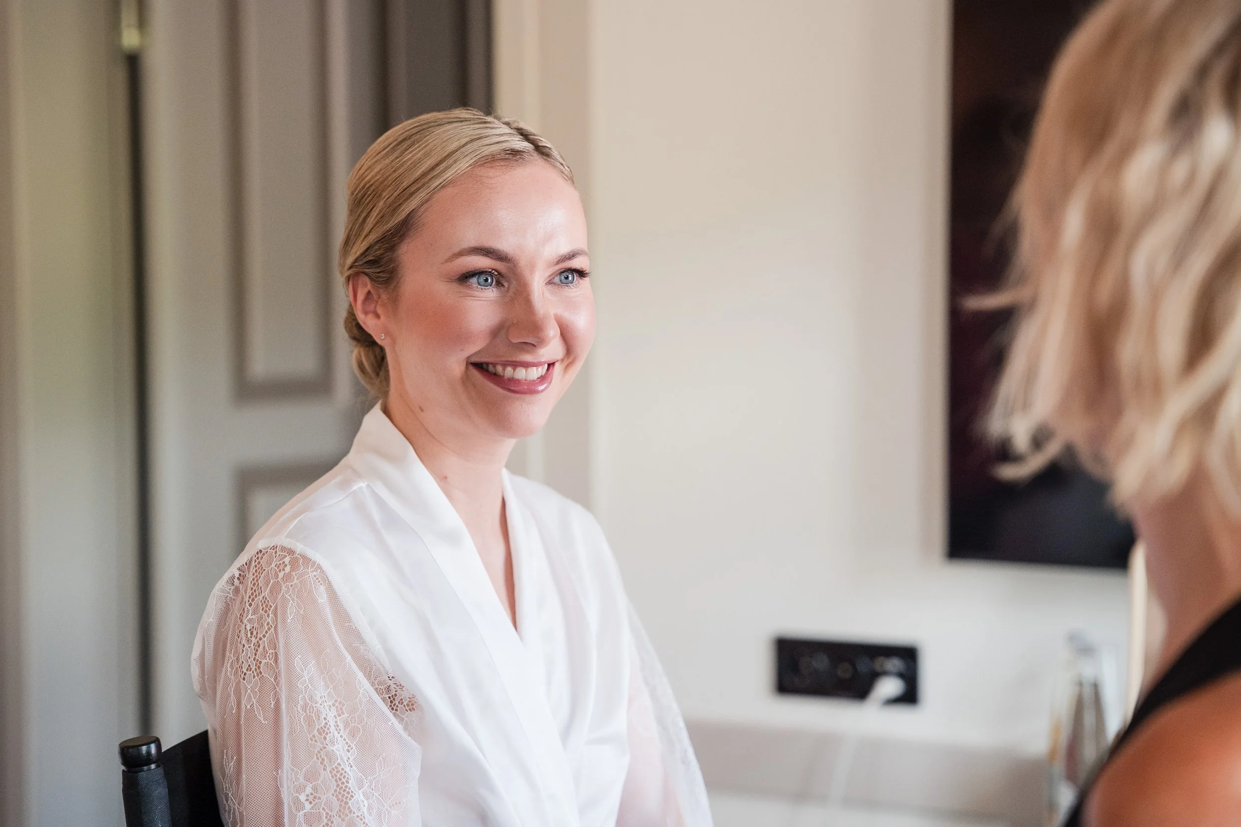 Zwei Frauen im Gespräch, eine lächelnde Frau mit blonden Haaren, die weiße Bluse trägt, und eine andere Frau, deren Gesicht nicht sichtbar ist.