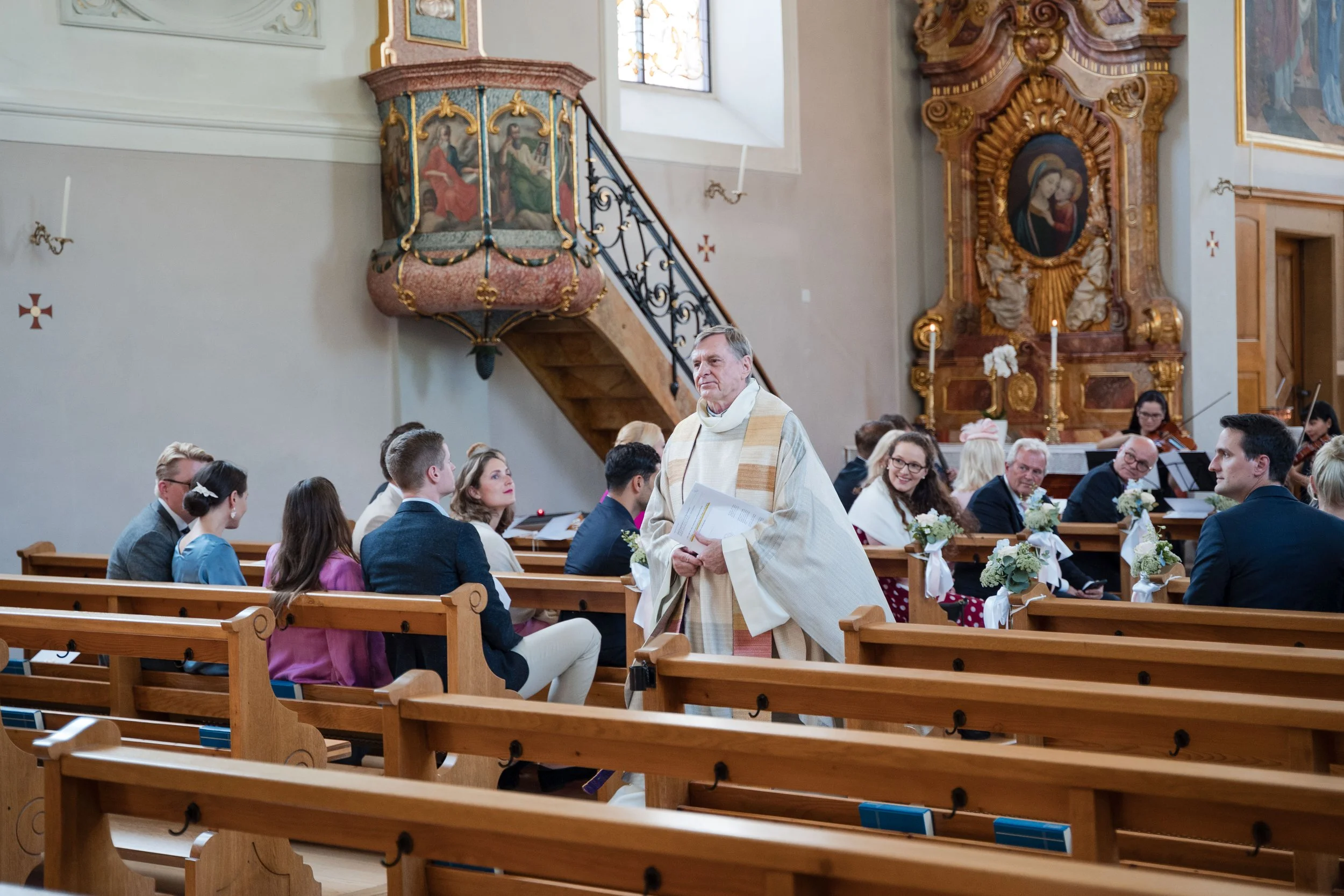 Priester in altem liturgischen Gewand spricht während einer kirchlichen Zeremonie in einer alten Kirche mit Holzstühlen, Dekorationen und einem Altar im Hintergrund, durch den der Chor spielt.