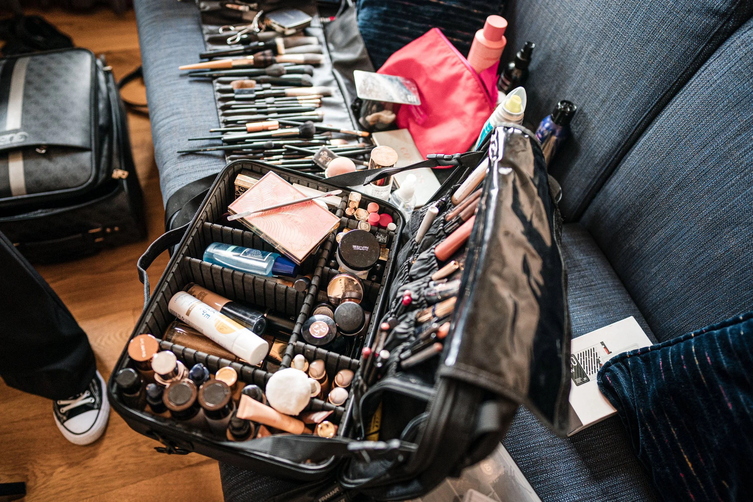 Offene Make-up-Beauty-Tasche auf einem Sofa mit vielen Make-up-Produkten, Pinseln und Utensilien, neben einem Rucksack und anderen Kosmetikartikeln