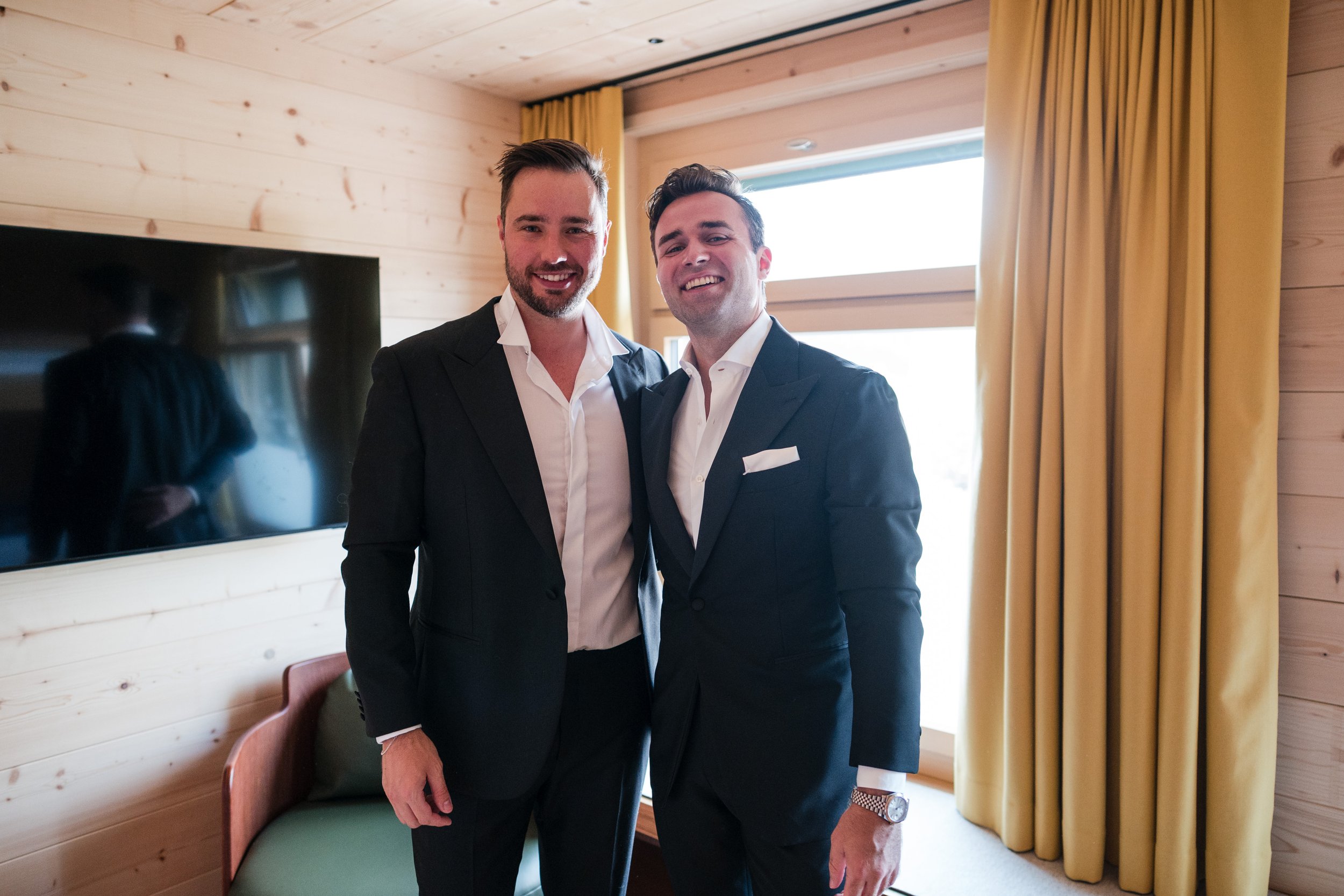 Zwei Männer in Anzügen stehen nebeneinander in einem Raum mit Holzvertäfelung, gelben Vorhängen und einem Fernseher an der Wand.