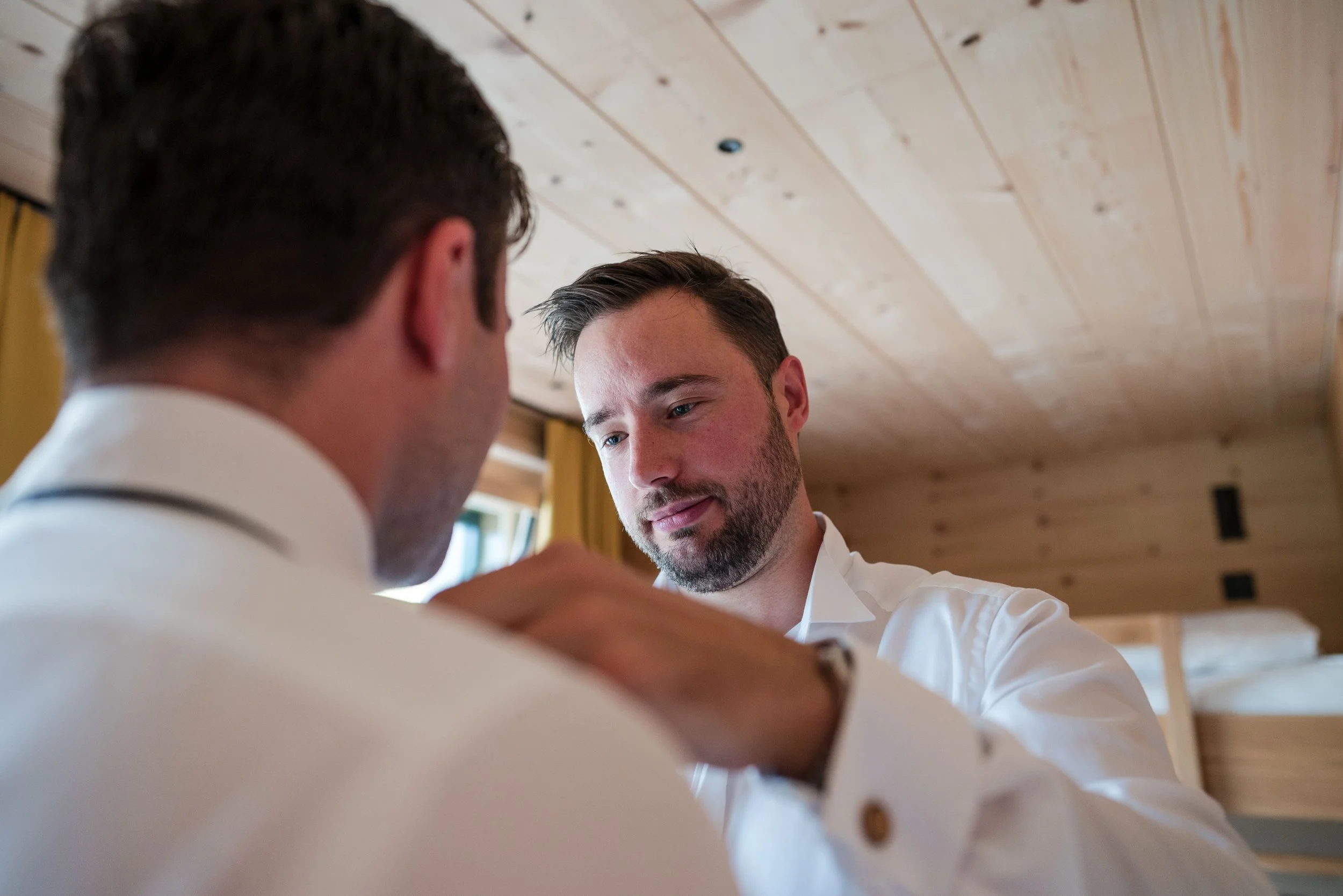 Zwei Männer in weißen Hemden, einer hilft dem anderen beim Anziehen, im Hintergrund eine Holzwand und ein Bett.